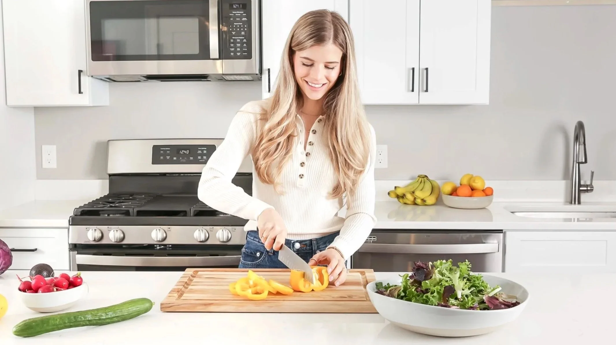A woman chopping yellow bell peppers in a modern kitchen with white cabinets, a stainless steel microwave, and a stove behind her. There are bowls of vegetables and fruit on the counter, including radishes, cucumbers, bananas, and apples.