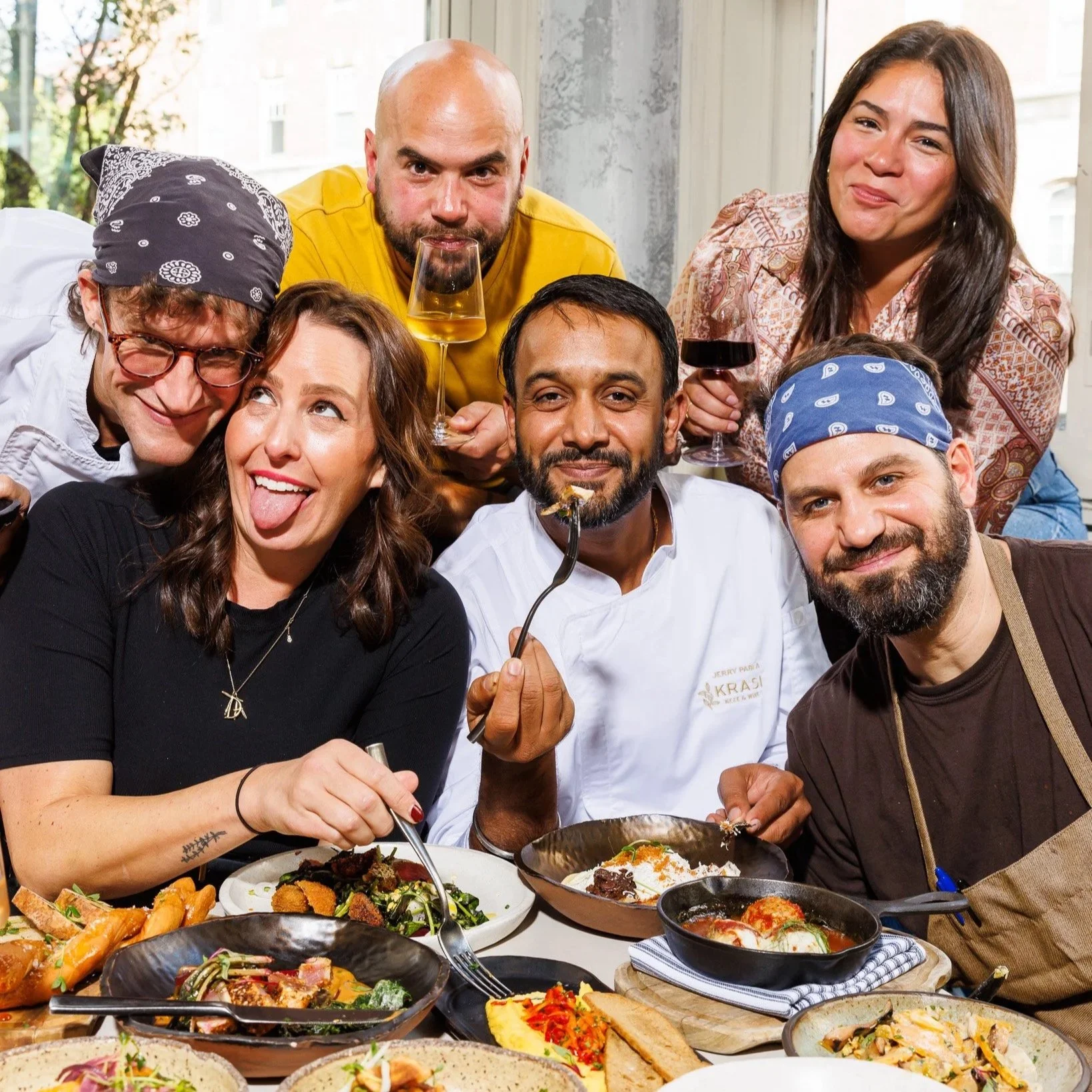 Group of six friends enjoying a meal together at a restaurant, with plates of food and drinks on the table.