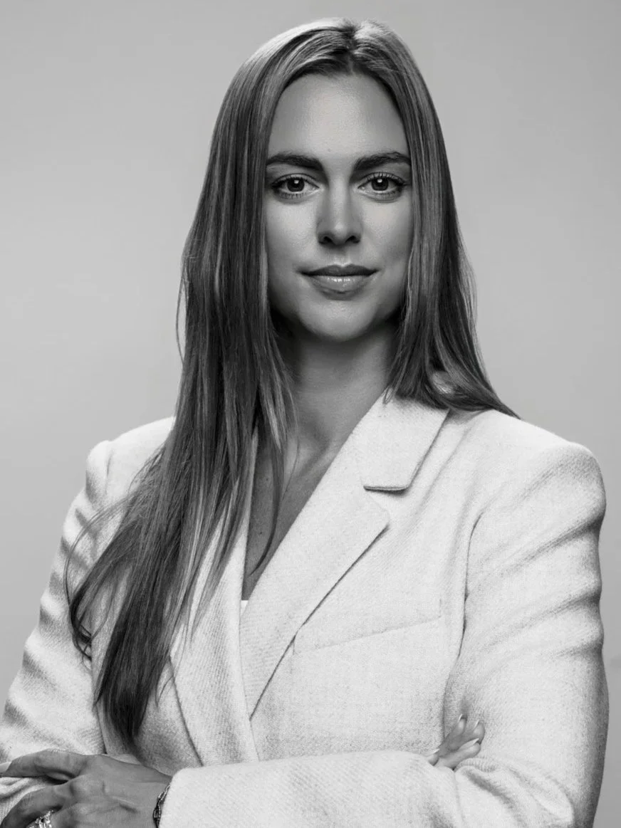 A black and white portrait of a woman with long, straight hair, wearing a light-colored blazer, standing with crossed arms.