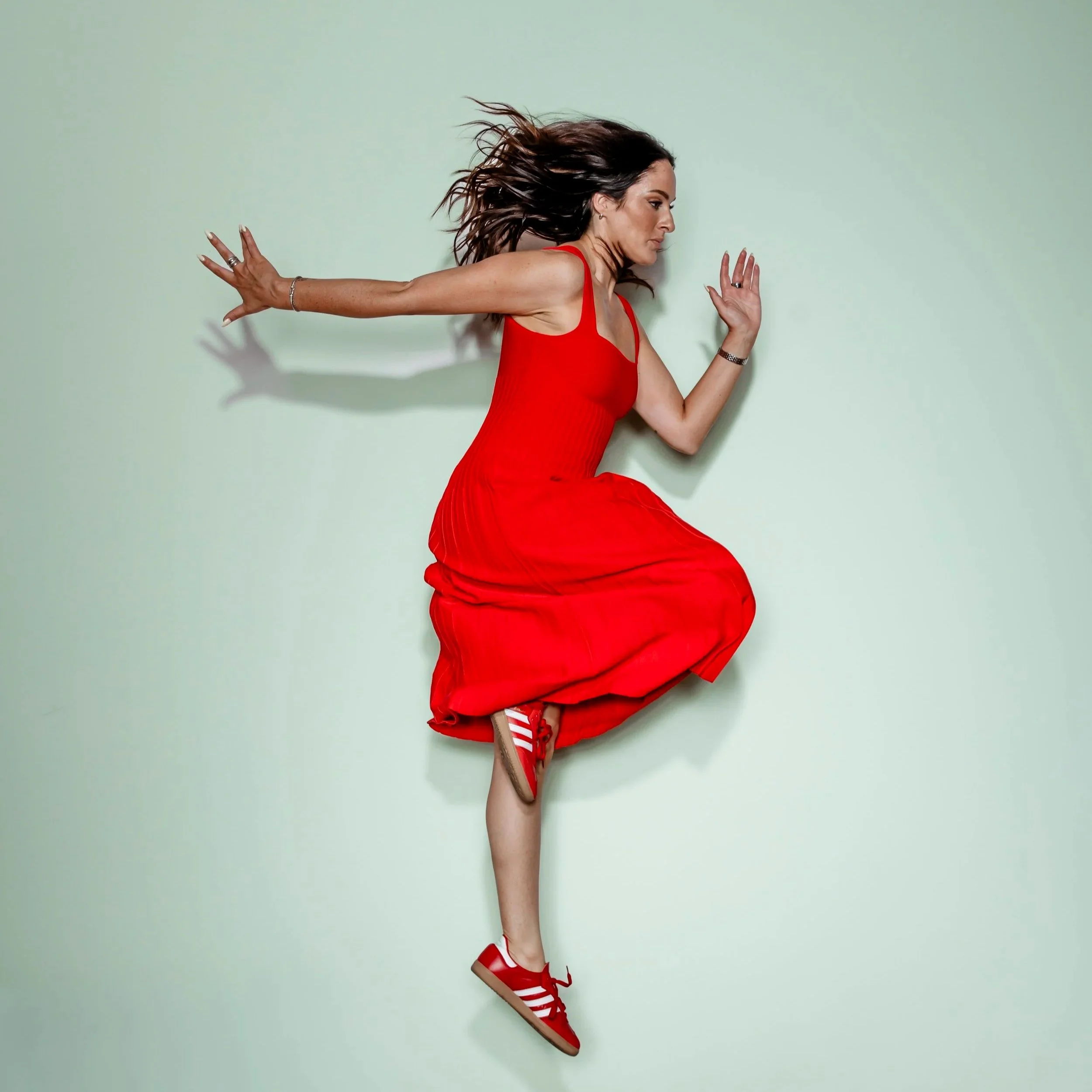 Woman in a red dress and red striped shoes leaning to the side with hair flowing, against a light green background.