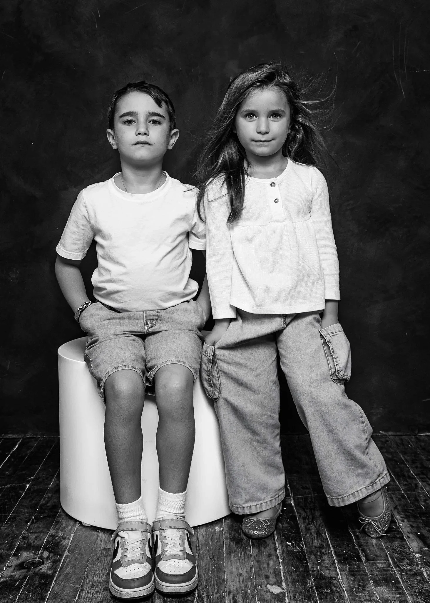 Black and white photograph of a boy and girl sitting side by side against a dark background. The boy is on the left, wearing a white T-shirt, shorts, and sneakers, with hands in pockets and a serious expression. The girl is on the right, wearing a lo