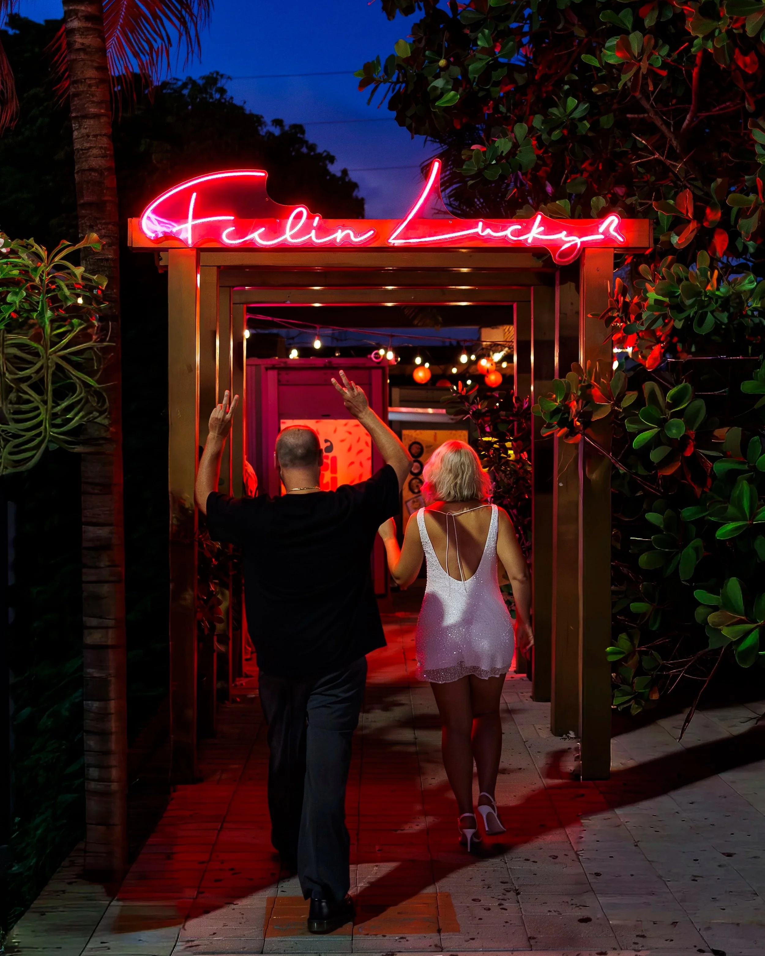 A man and a woman walking into a night event through an entrance decorated with a pink neon sign that says 'Falin Lucky'. The woman is wearing a shiny white dress with high heels, and the man is wearing dark pants and a black shirt. The scene is illu