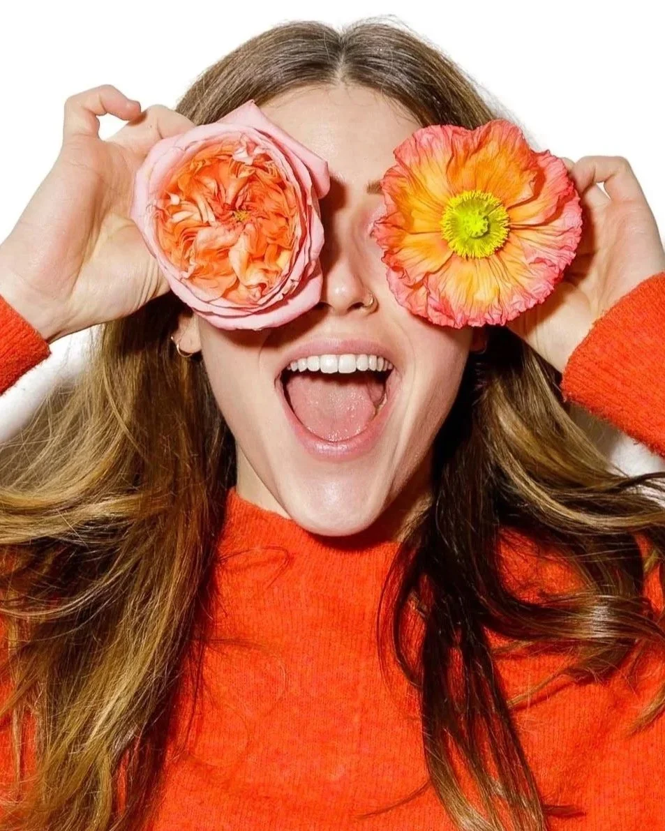 A woman with long, wavy red hair holding two colorful flowers over her eyes, smiling widely with her mouth open, wearing an orange sweater.