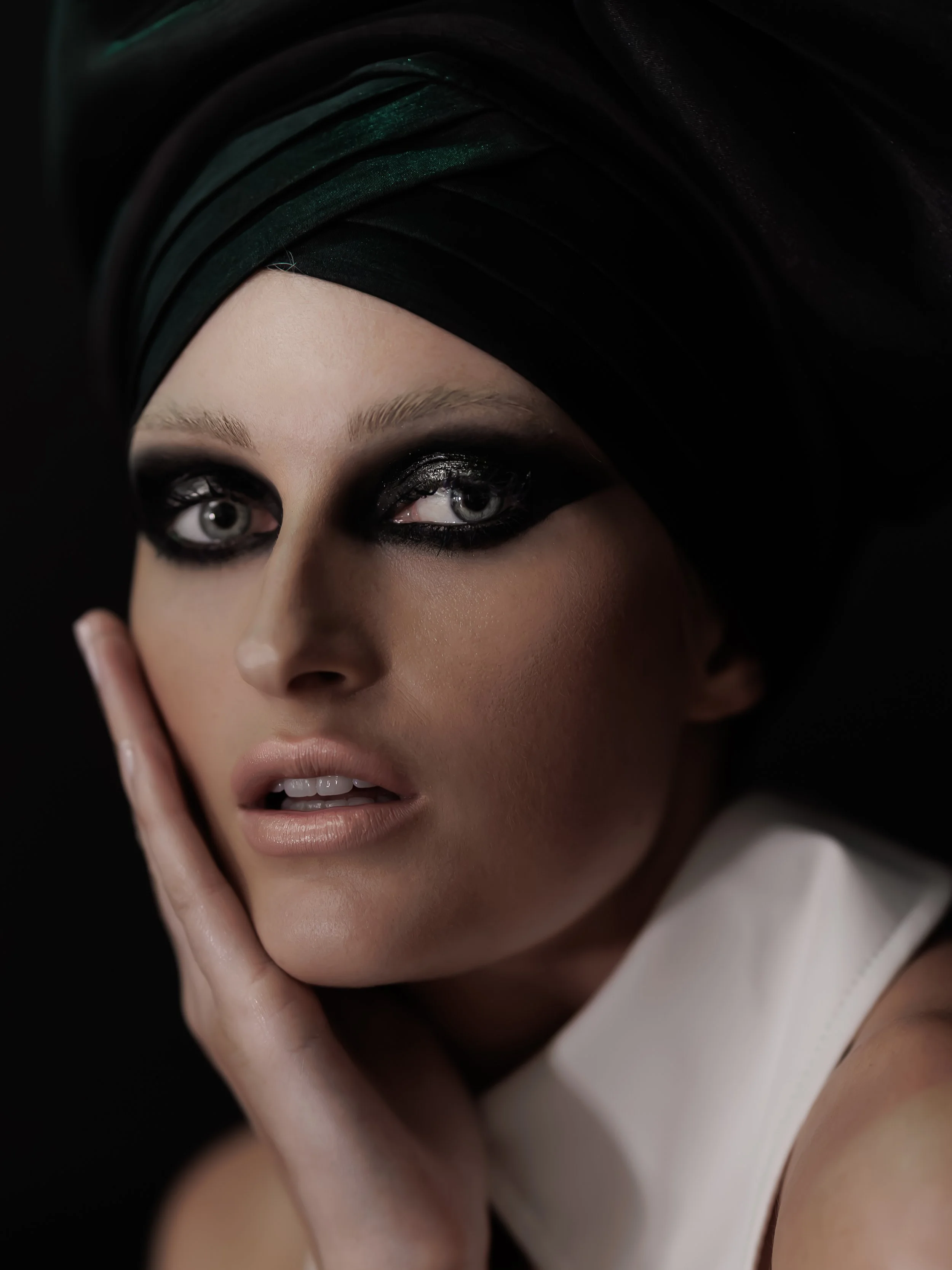 Close-up of a woman's face with bold black eye makeup, wearing a black headscarf with green sheen, resting her chin on her hand, against a black background.