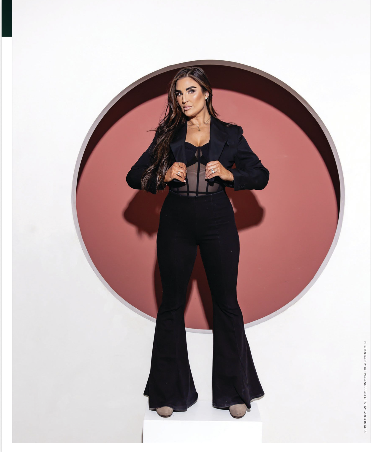 A woman with long dark hair standing in front of a circular red background, dressed in a black sheer top, black pants, and beige shoes.
