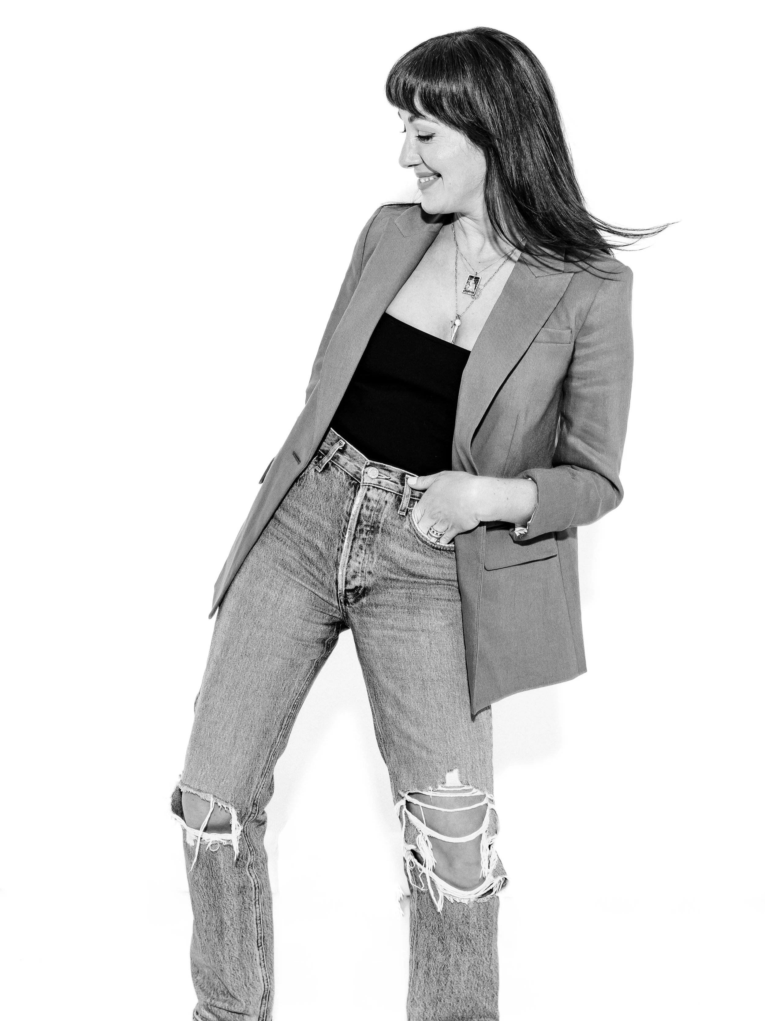 A woman with shoulder-length dark hair wearing a blazer, black top, ripped jeans, and layered necklaces, smiling and looking down.