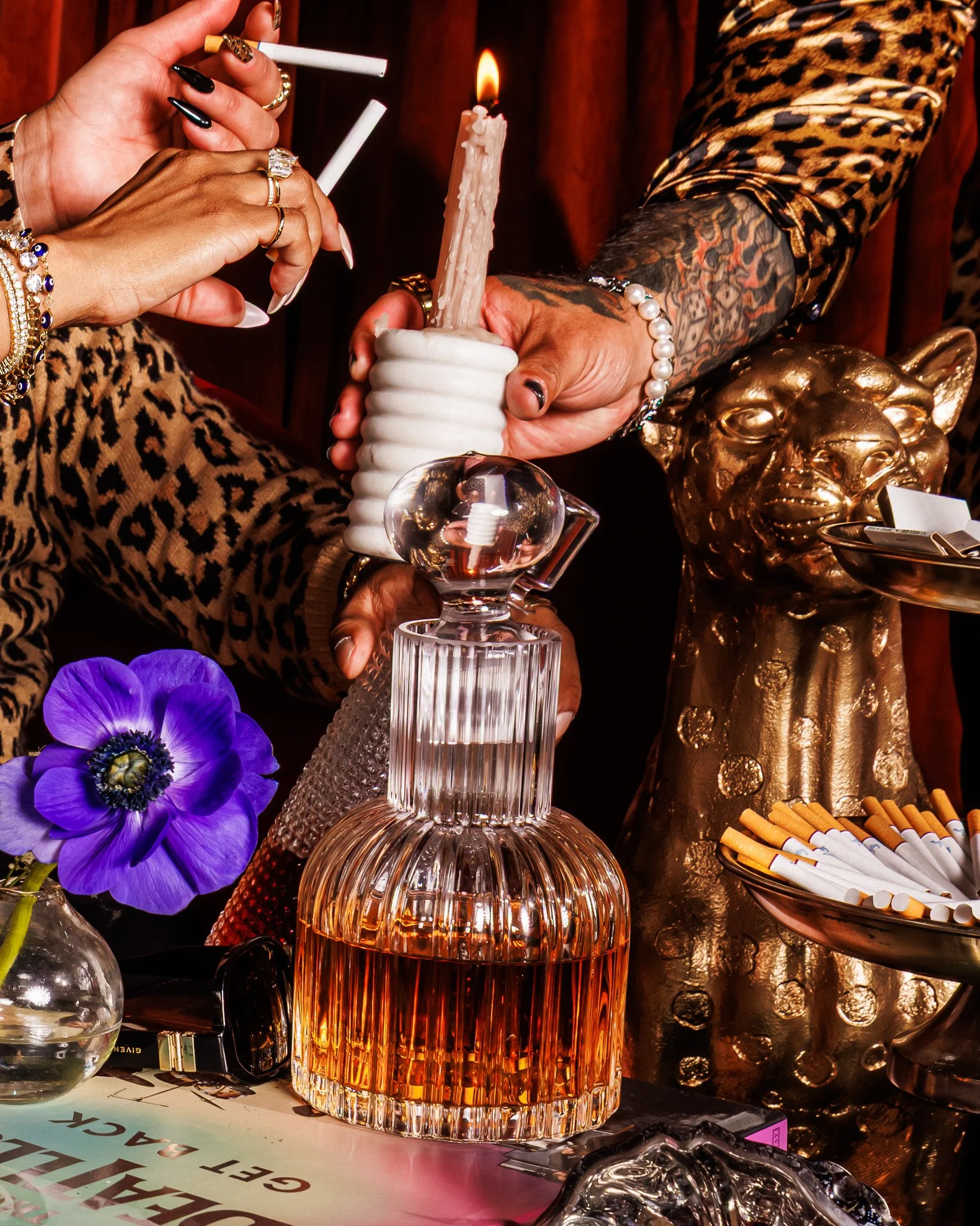 Two people holding a lit candle over a glass decanter on a table with a purple flower, cigarettes, and a golden lion statue.