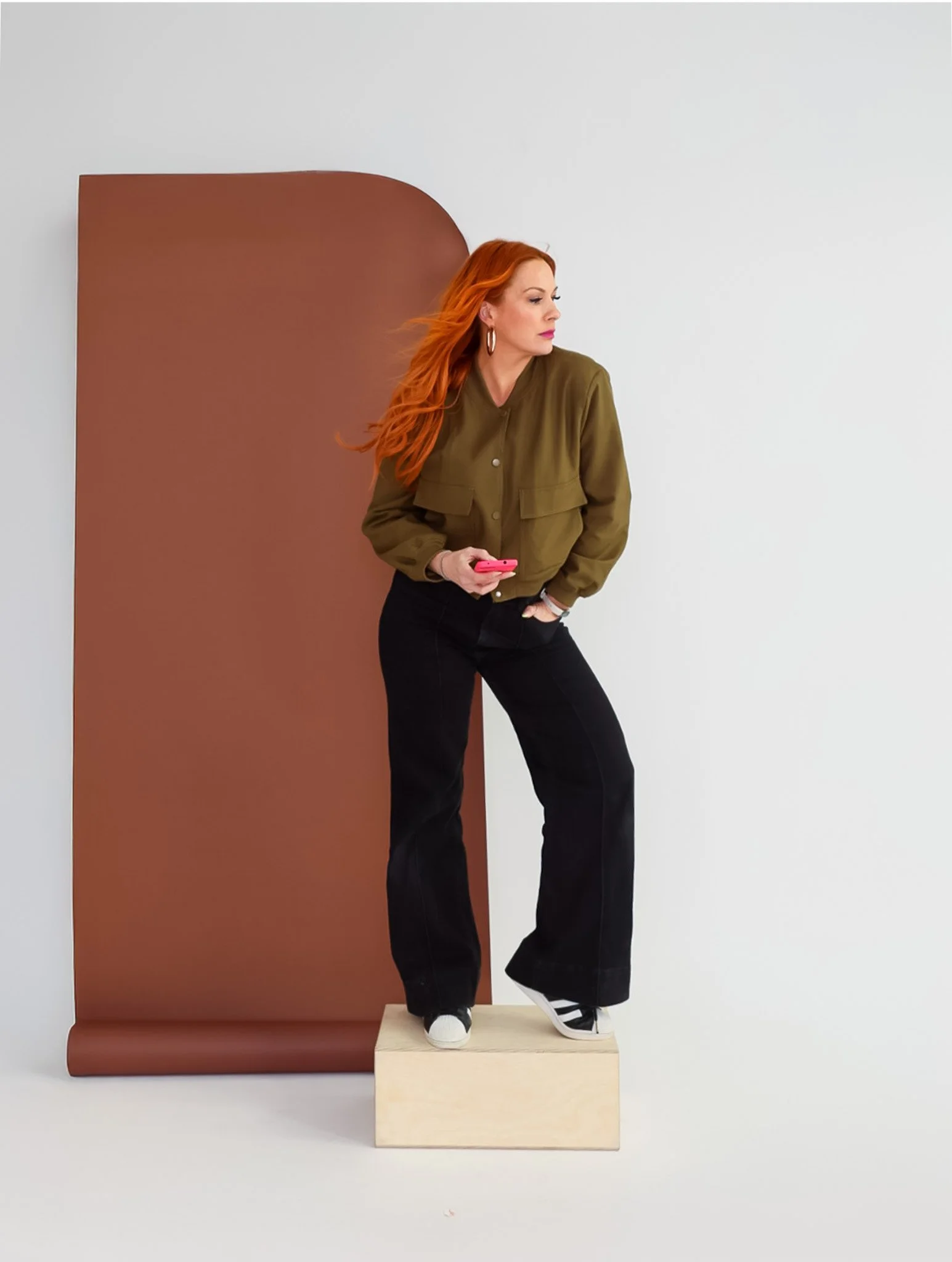 A woman with long red hair standing on a wooden block, wearing black pants, a green jacket, and white sneakers, holding a pink phone, with a brown backdrop behind her on the left side and a plain white wall on the right.