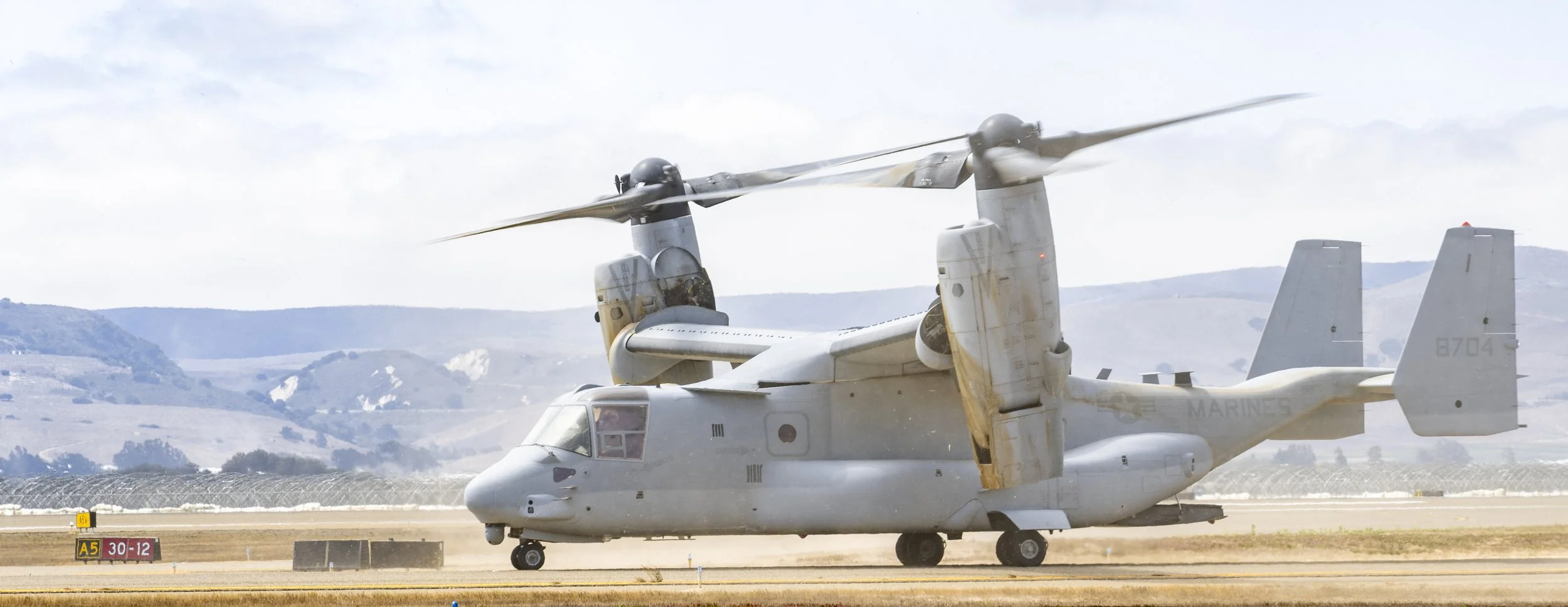 A military tiltrotor aircraft on the runway with its rotors in a vertical position and propellers spinning, preparing for takeoff, with scenic hills in the background under a cloudy sky.