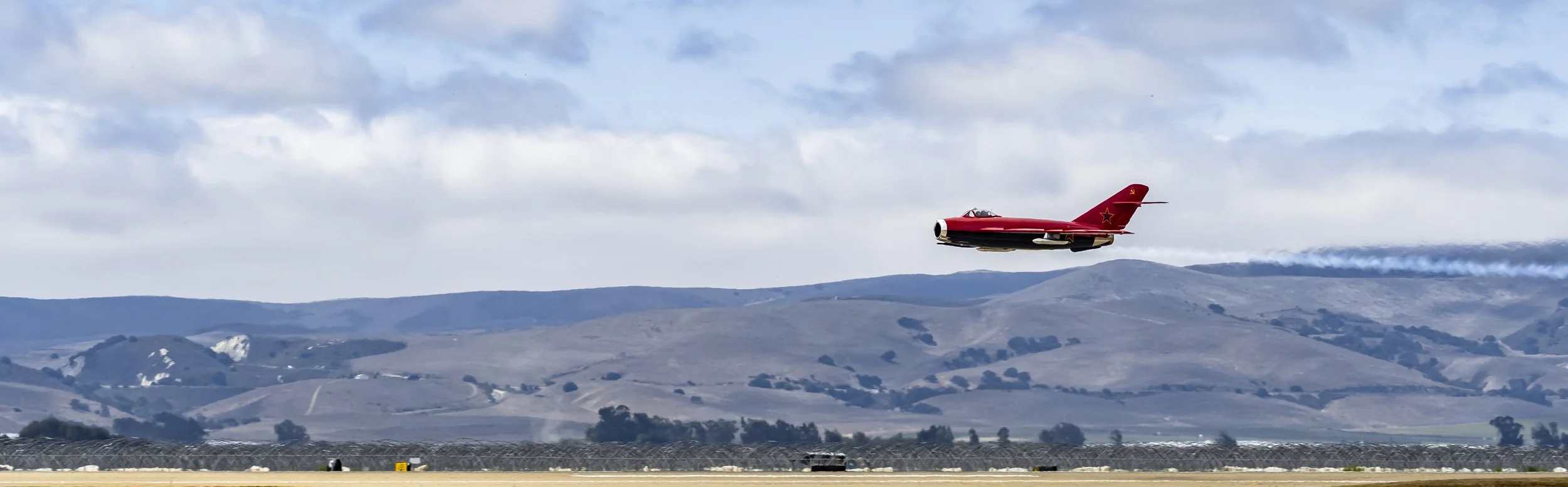 A red jet airplane flying low over a mountain range on a cloudy day.