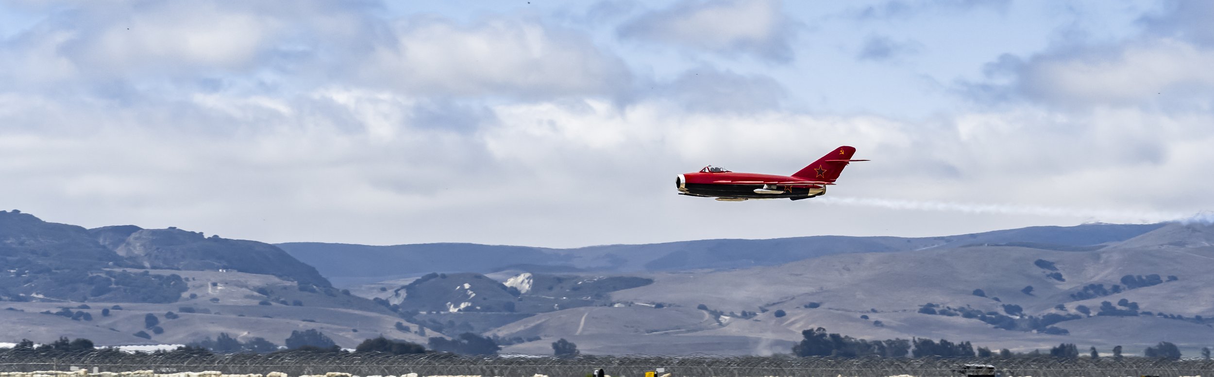 A red jet aircraft flying low over a landscape with rolling hills and partly cloudy sky.