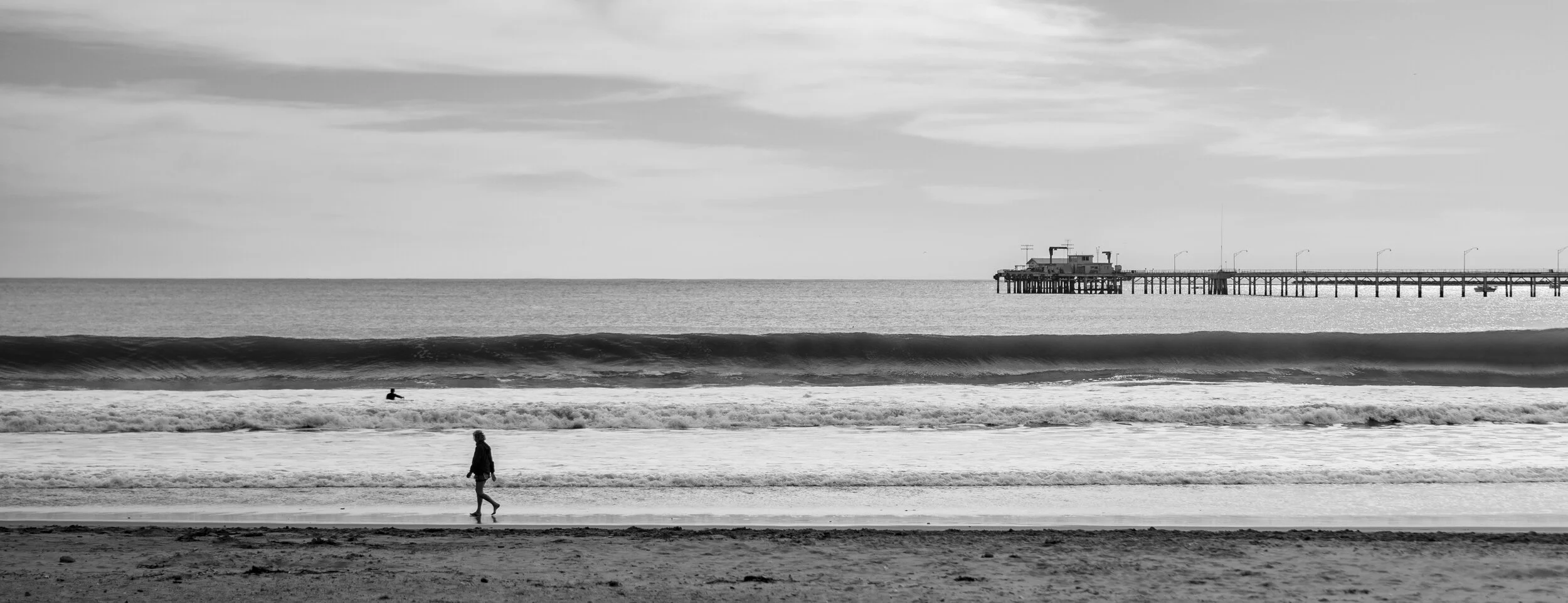Black and white photo of a beach with a pier extending into the ocean. A person is walking along the shoreline, and a surfer is in the water near the waves.