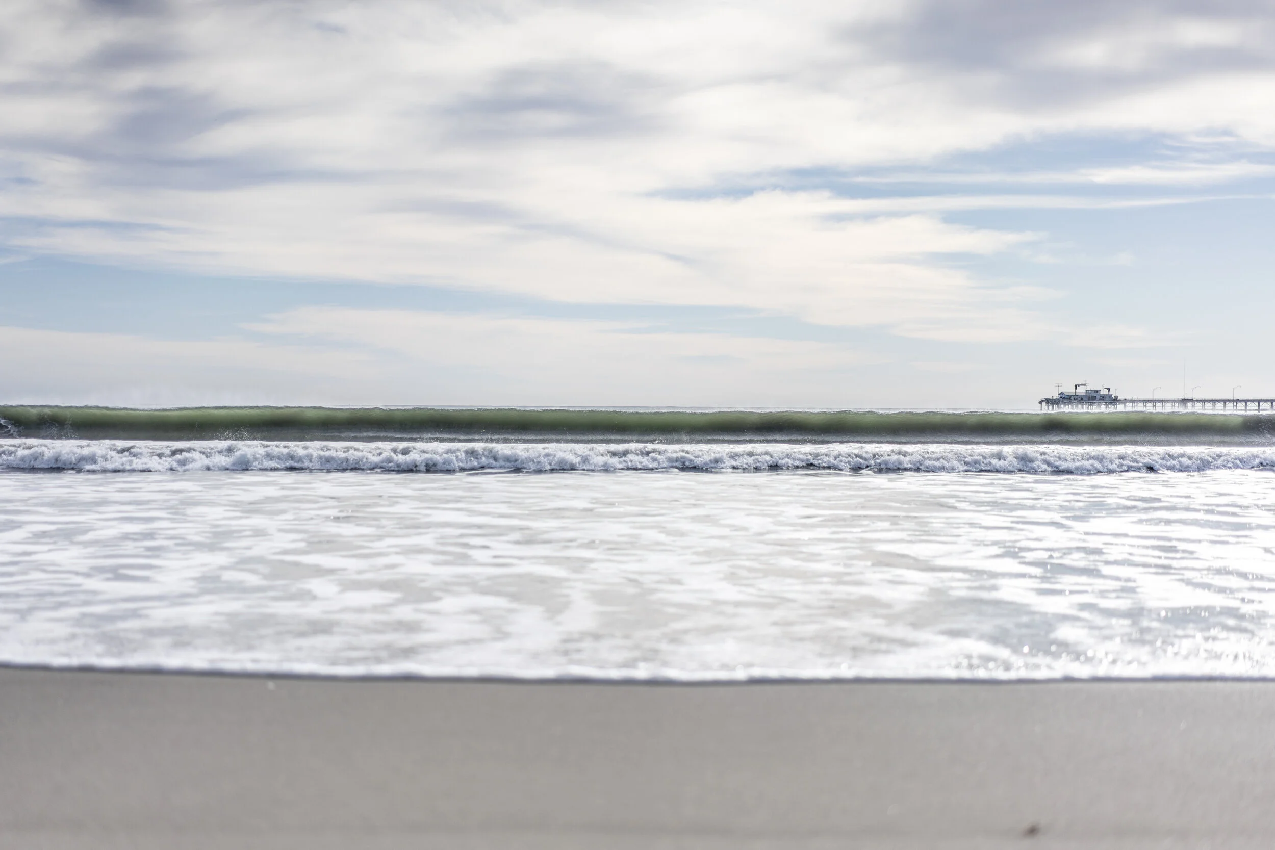 View of a pier extending into the ocean with waves and a cloudy sky.