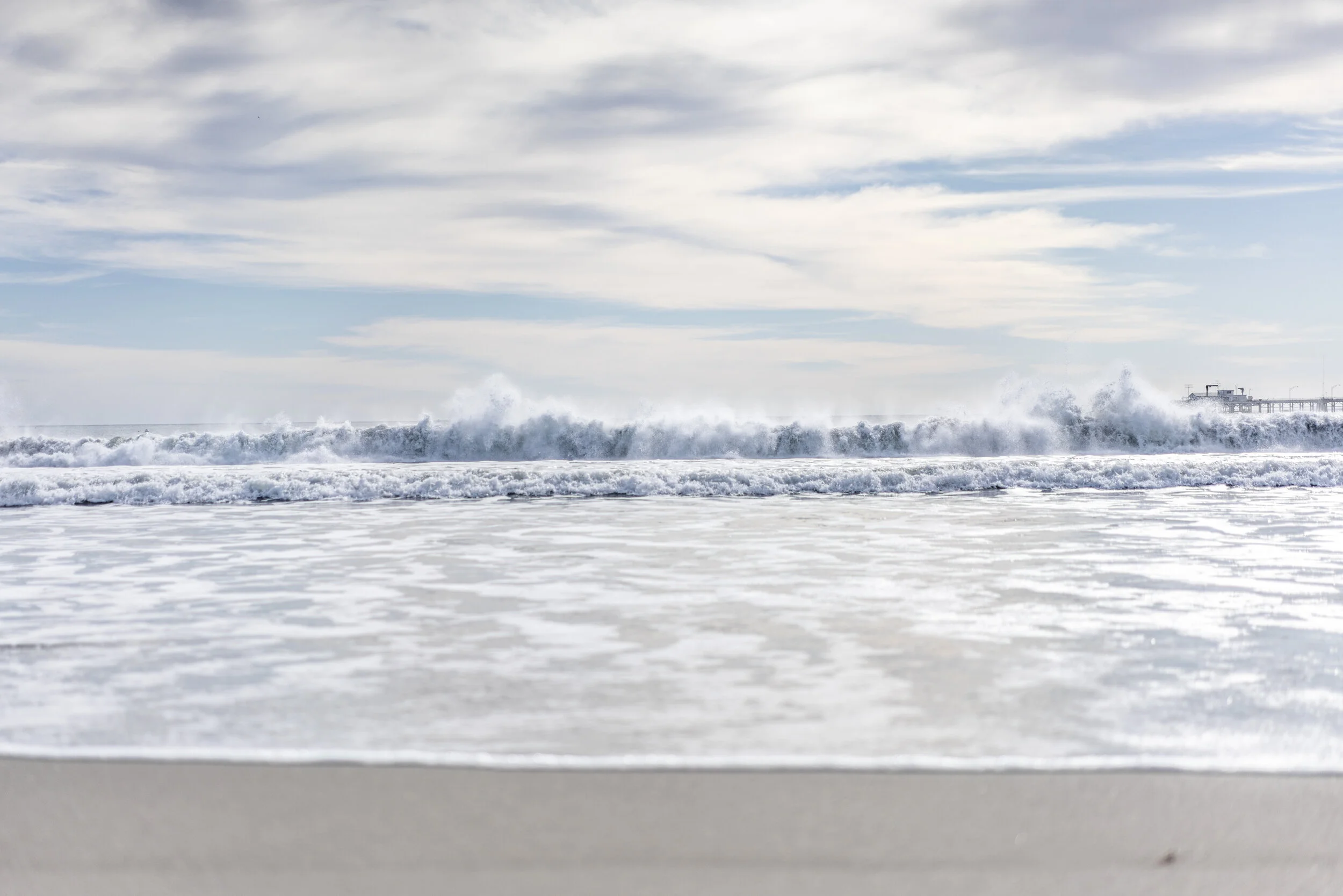 Ocean waves crashing against the shoreline with a pier visible in the background under a cloudy sky.
