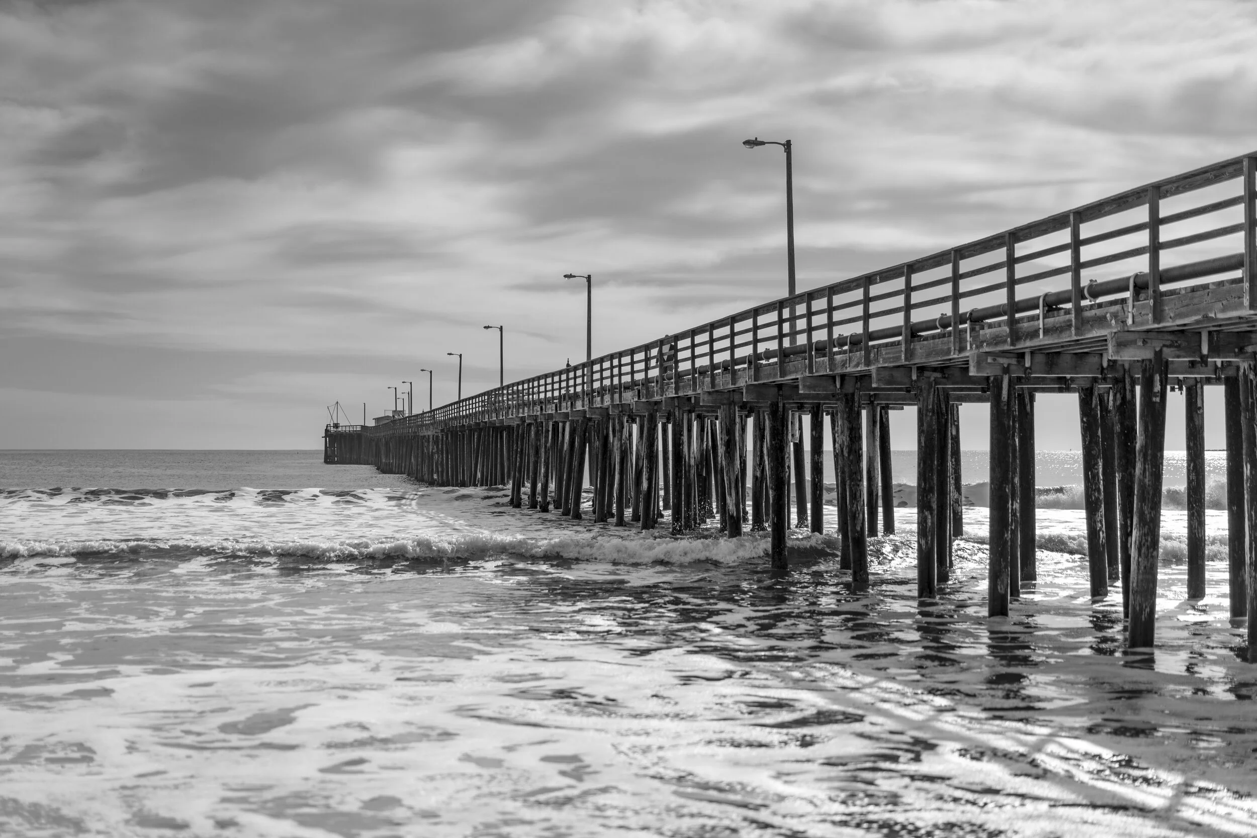 A black and white photograph of a wooden pier extending into the ocean with multiple street lamps along its length, and the sky filled with scattered clouds.