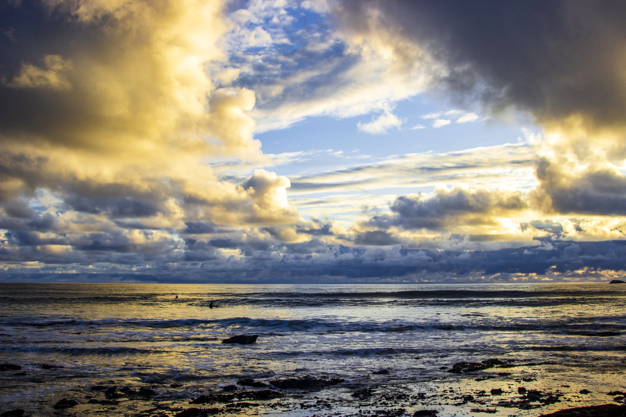 A sunset over the ocean with a sky filled with dark and yellow clouds, some appearing to be illuminated by the setting sun, and a few small waves and rocks in the foreground.