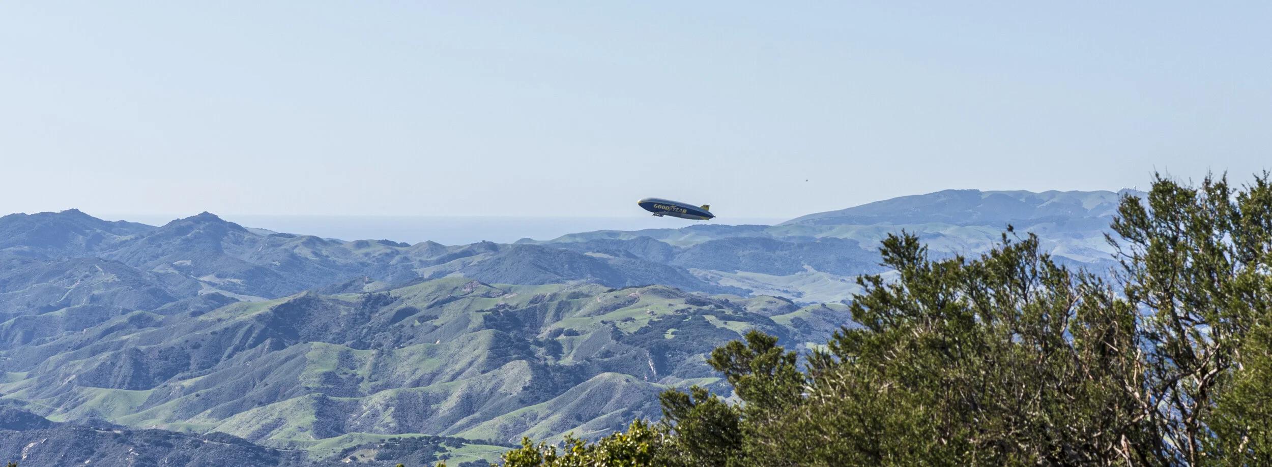 A blimp flying over green hilly landscape with trees in the foreground, blue sky in the background.