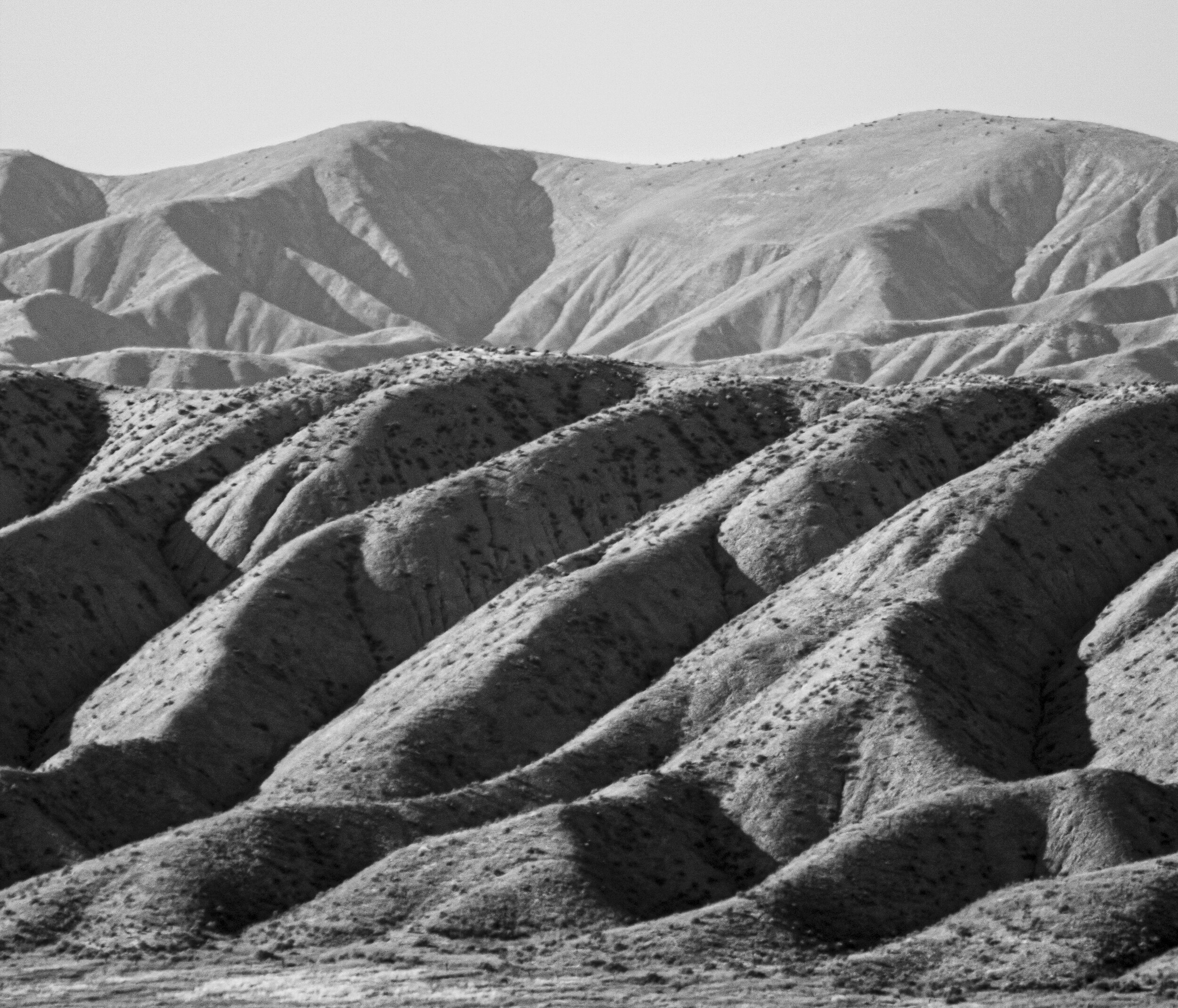 Black and white photograph of rugged, eroded hills and mountains with deep valleys, showing layered sedimentary formations.