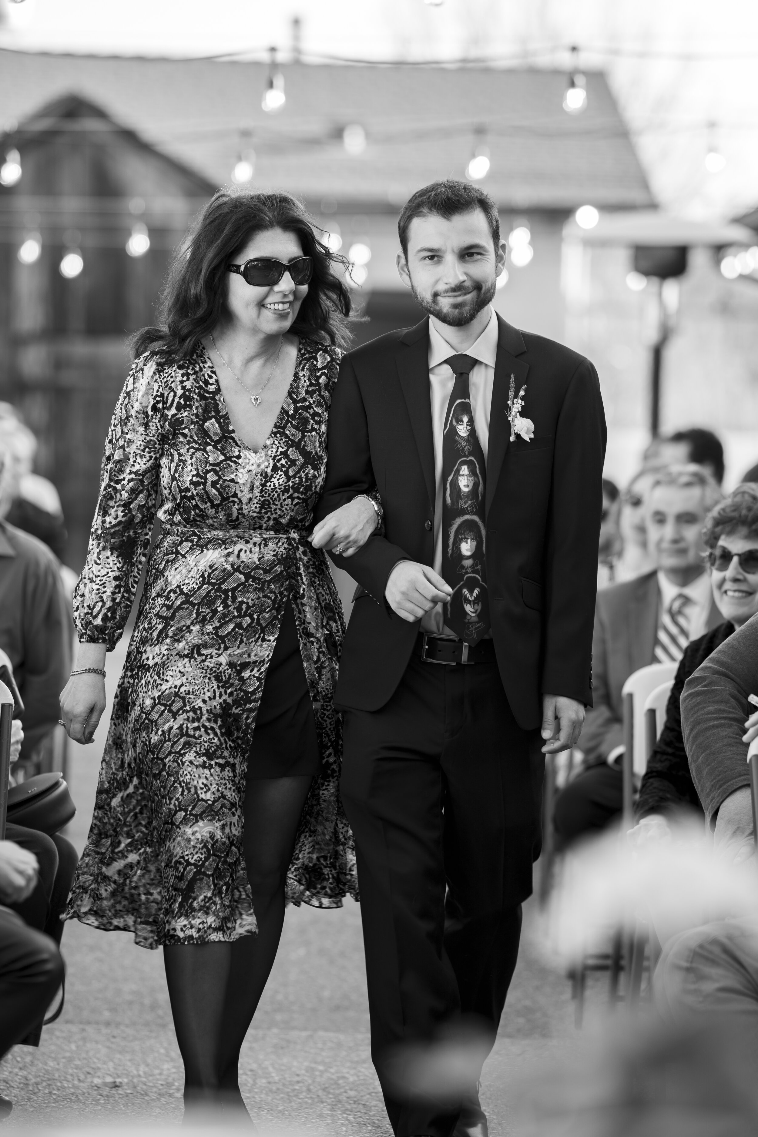 A black and white photo of a young man in a suit and tie, smiling, walking arm-in-arm with a woman wearing sunglasses, a patterned dress, and a heart necklace, at an outdoor event with chairs and people seated in the background, strung lights overhea