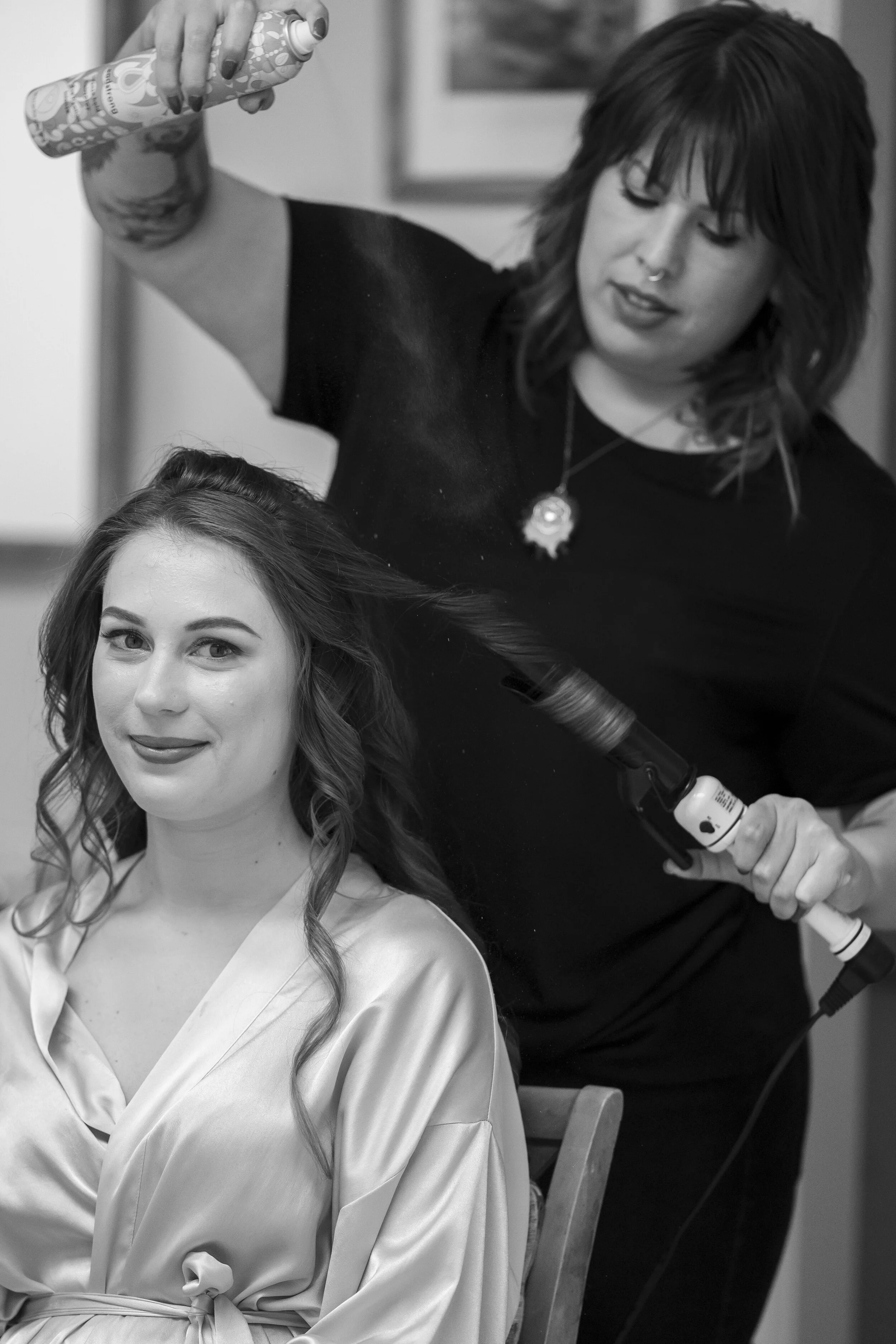 A woman with wavy hair and a satin robe is sitting while a hairstylist uses a curling iron on her hair.
