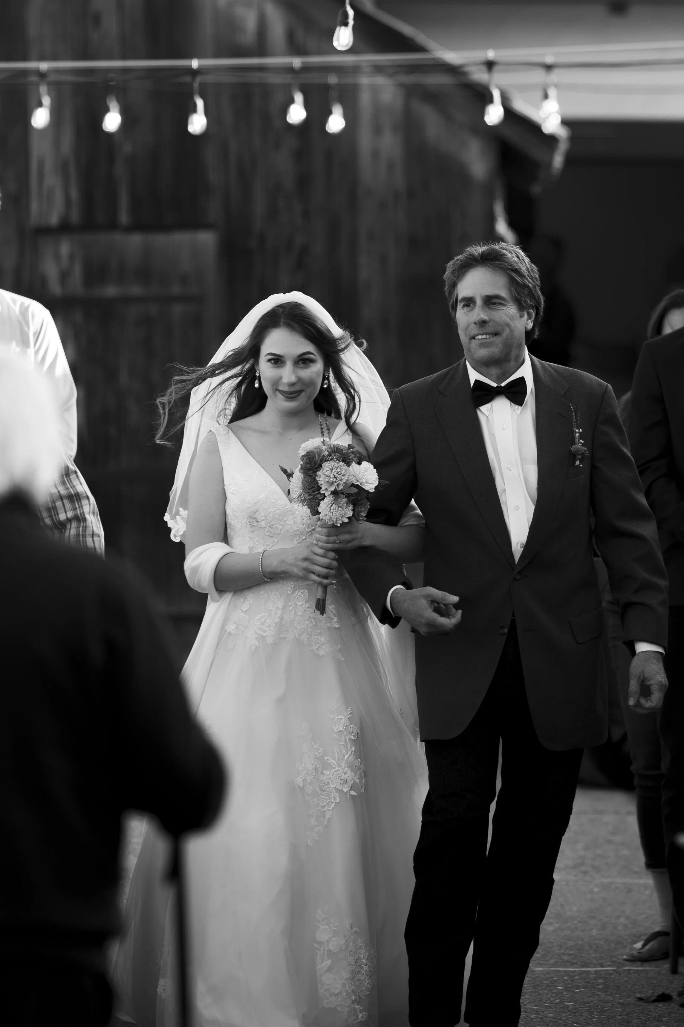A bride in a white wedding dress holding a bouquet of flowers walks arm in arm with a man in a tuxedo during a wedding ceremony in a rustic indoor setting.