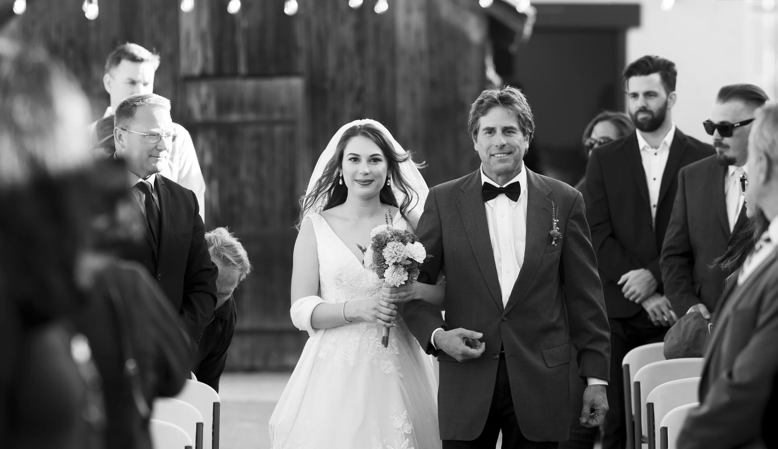 Black and white photo of a bride in a wedding dress holding a bouquet, walking down the aisle with a man in a suit. Several guests are on either side, some standing and some seated, watching her.