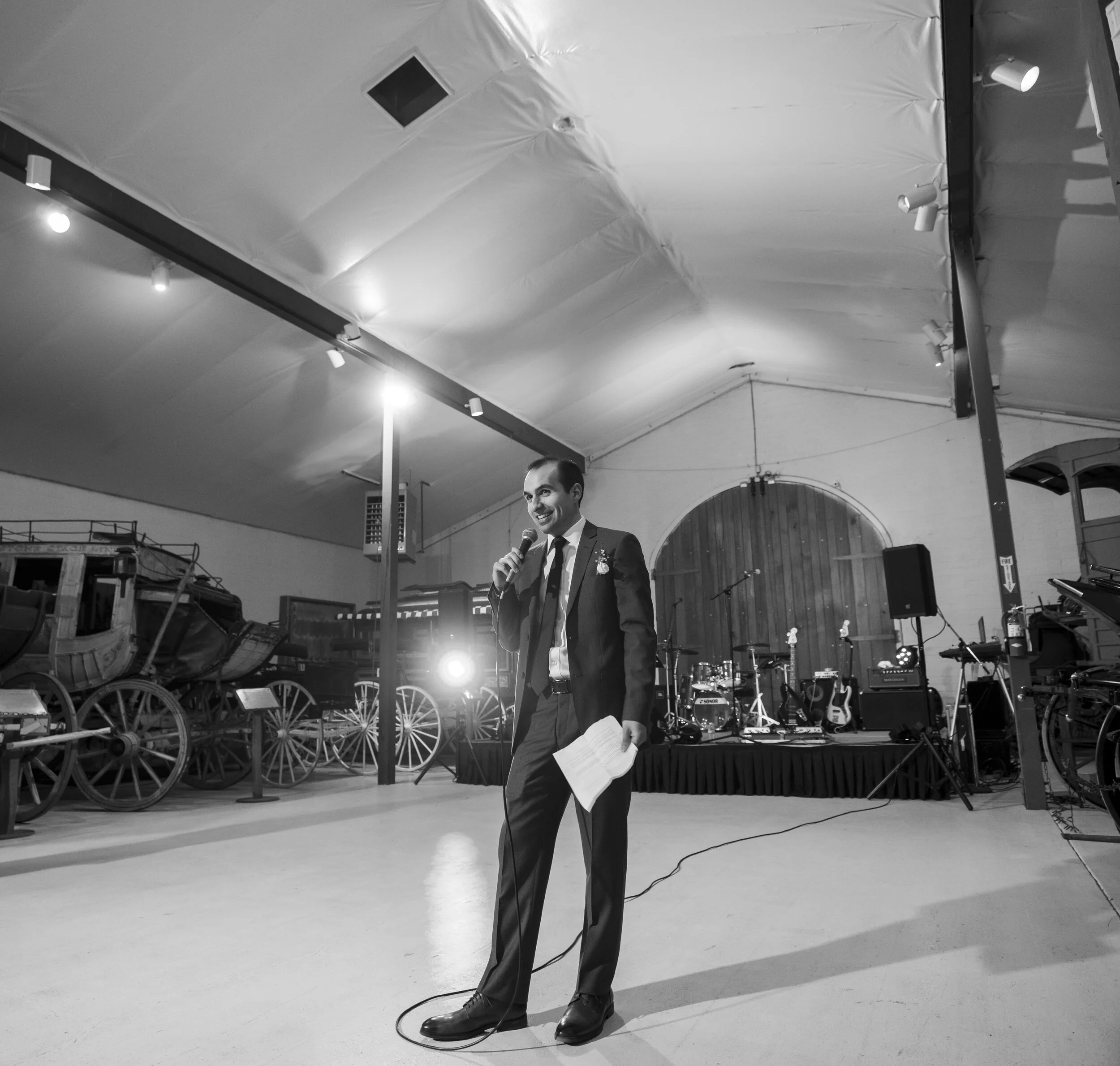 A man in a suit holding a microphone and a sheet of paper, standing on a stage with musical instruments in the background, under a curved ceiling with spotlights and ceiling tiles.