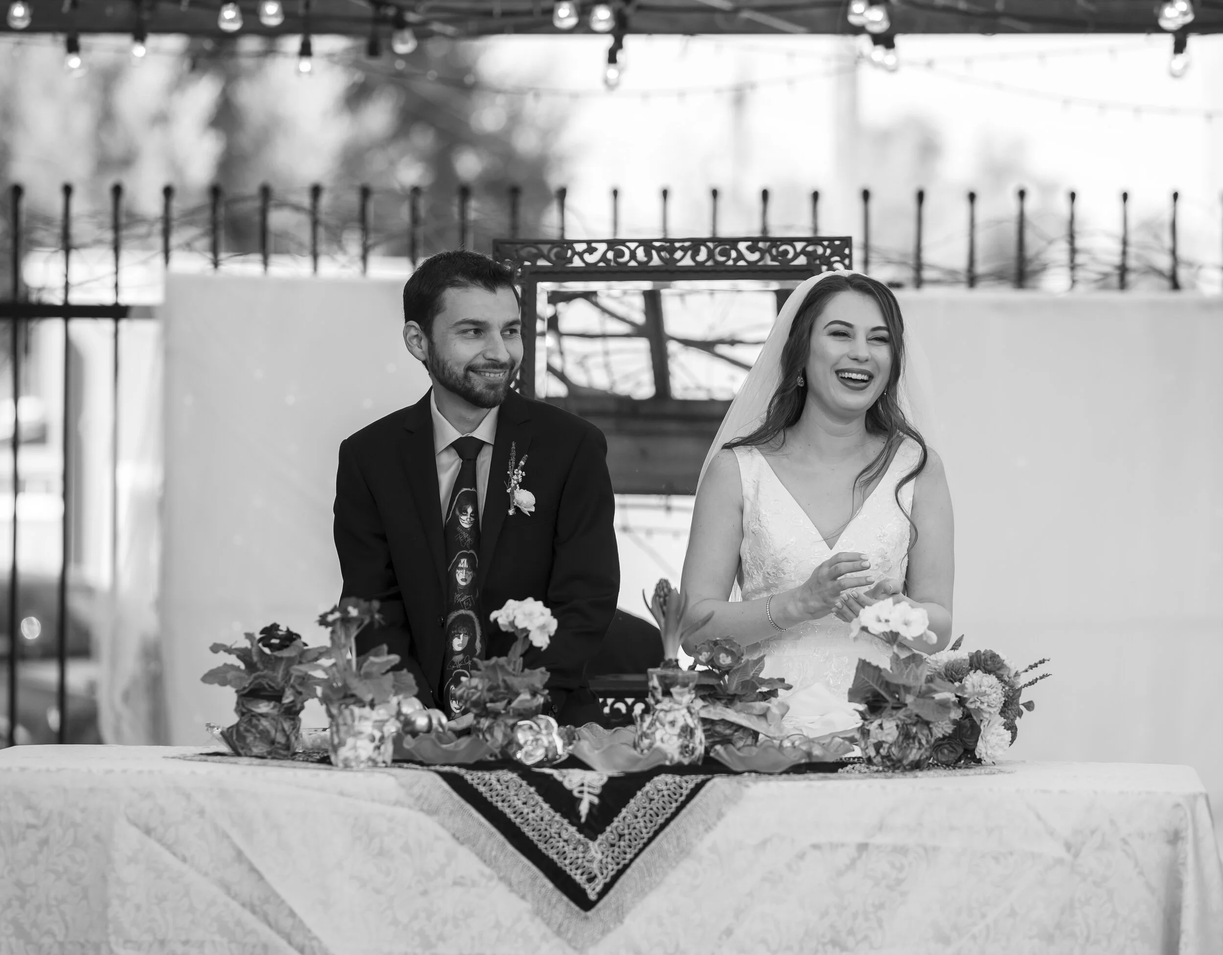 A black and white photo of a man and woman dressed in wedding attire sitting at a table decorated with flowers, during a wedding ceremony indoors.