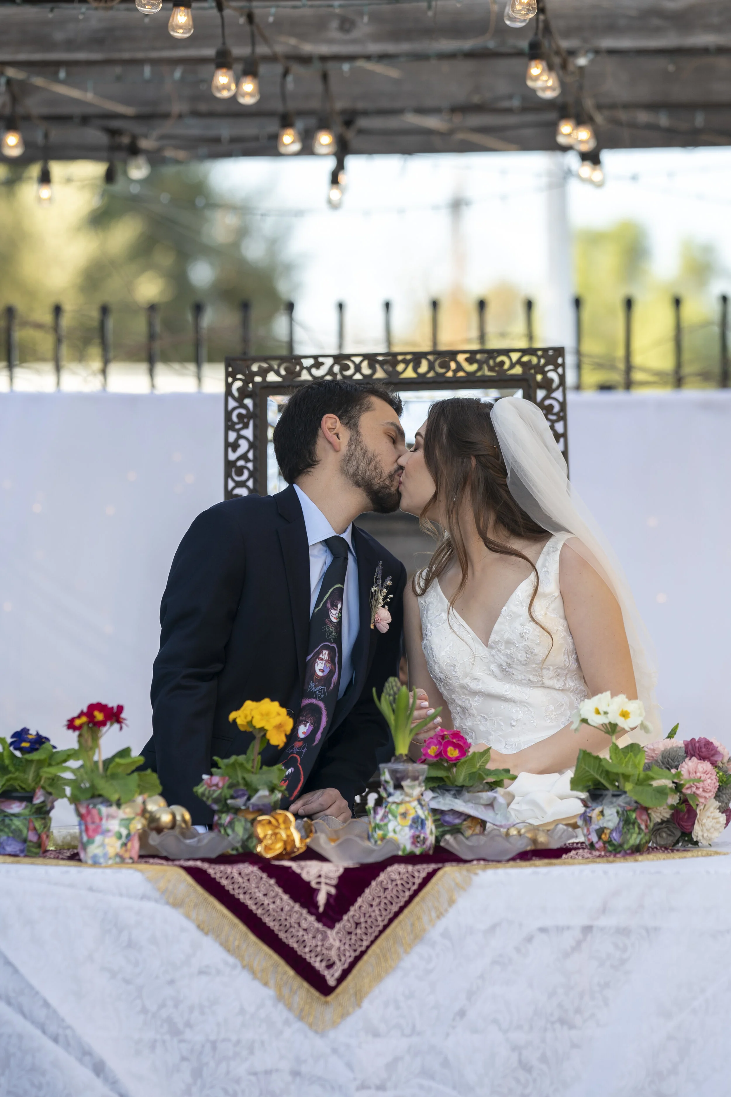 A bride and groom kiss at their wedding reception, standing behind a decorated table with colorful flowers. The bride wears a white wedding gown and veil, and the groom is dressed in a dark suit with a tie featuring images of faces. Overhead string l