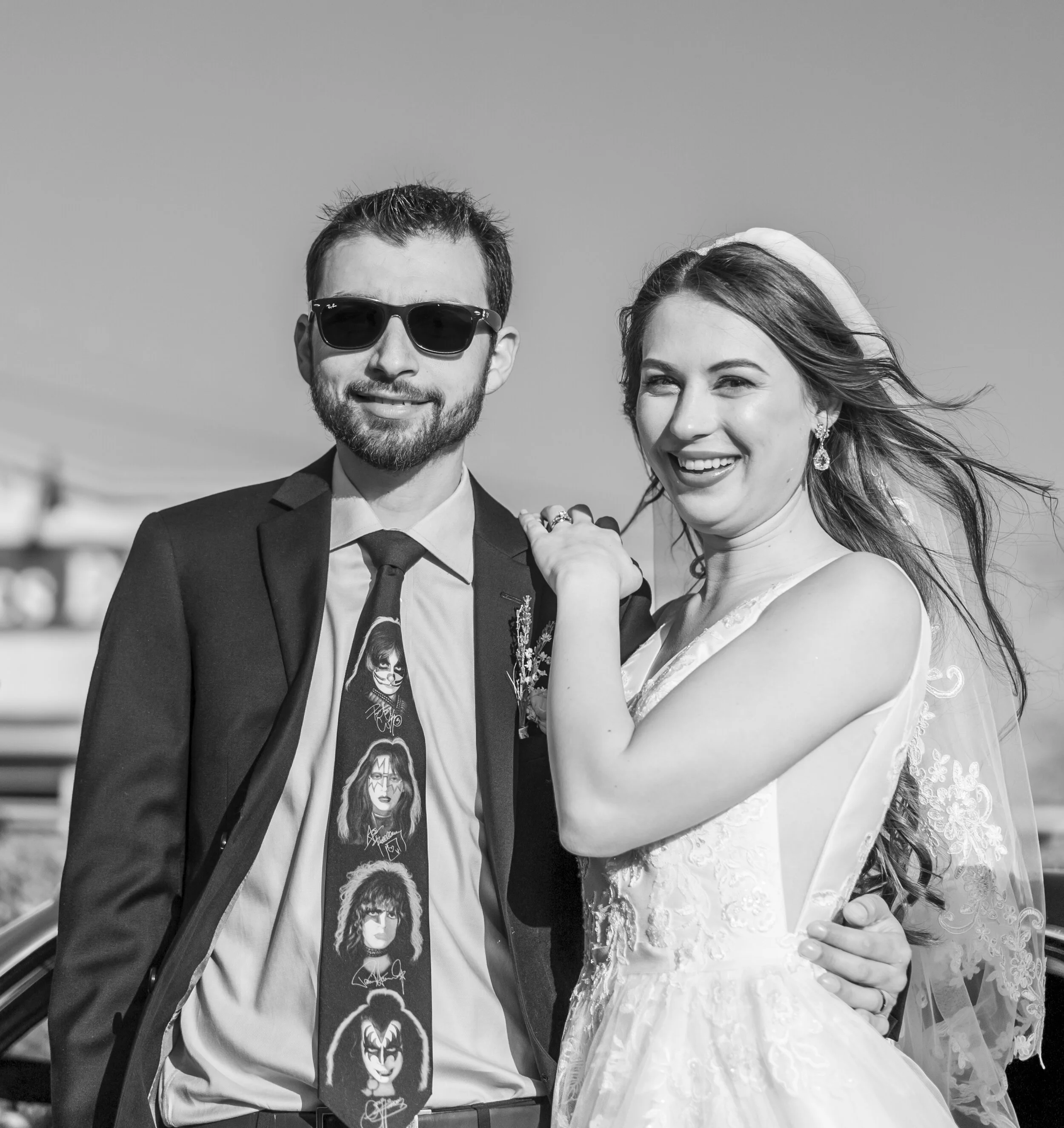 Black and white photo of a smiling man wearing sunglasses and a tuxedo, standing next to a smiling woman in a wedding dress and veil. The woman has earrings and is holding his hand.