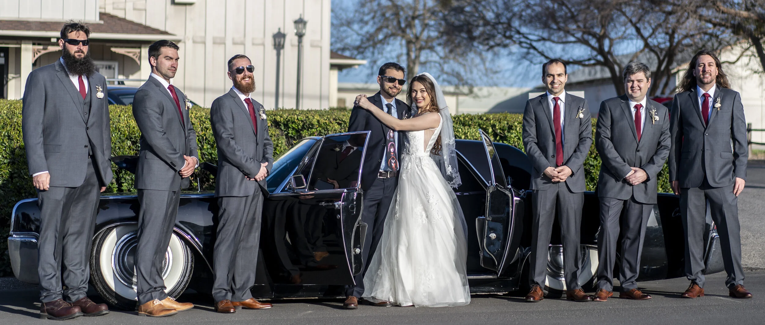 Wedding party standing beside a vintage black car with open doors. The bride and groom are in the center, hugging with the bride in a white wedding dress and the groom in a dark suit. Four groomsmen in matching gray suits with red ties and boutonnier