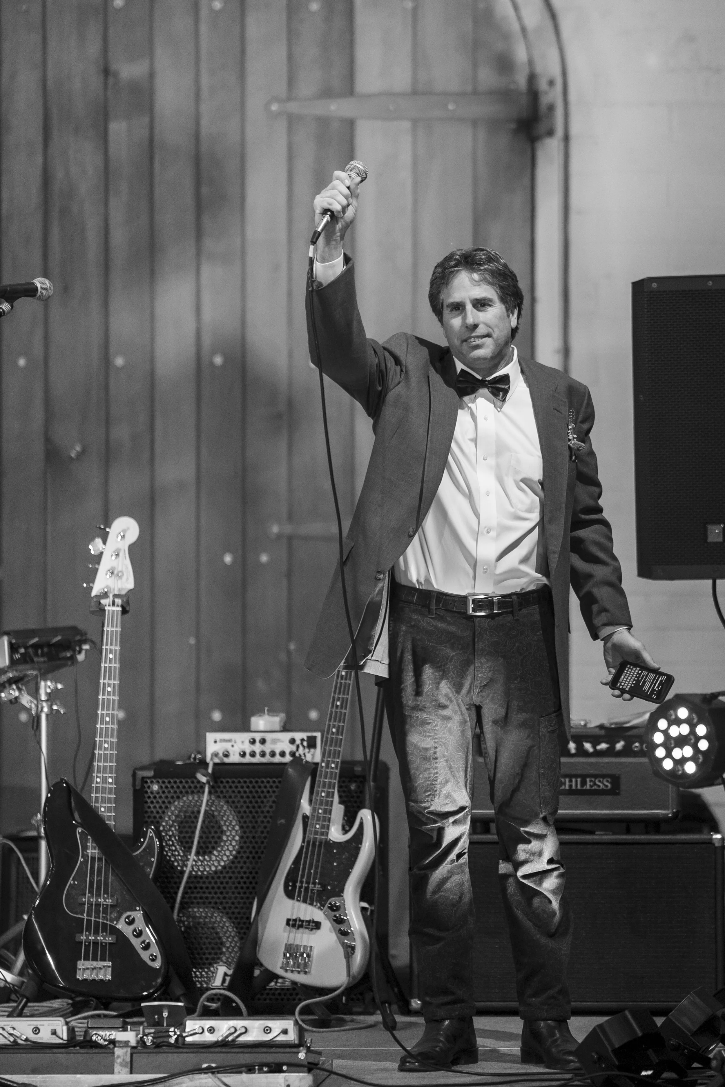 A man in formal attire, including a tuxedo jacket and bow tie, holding a microphone and standing on stage surrounded by musical equipment, guitars, and amplifiers, with a wooden wall in the background.