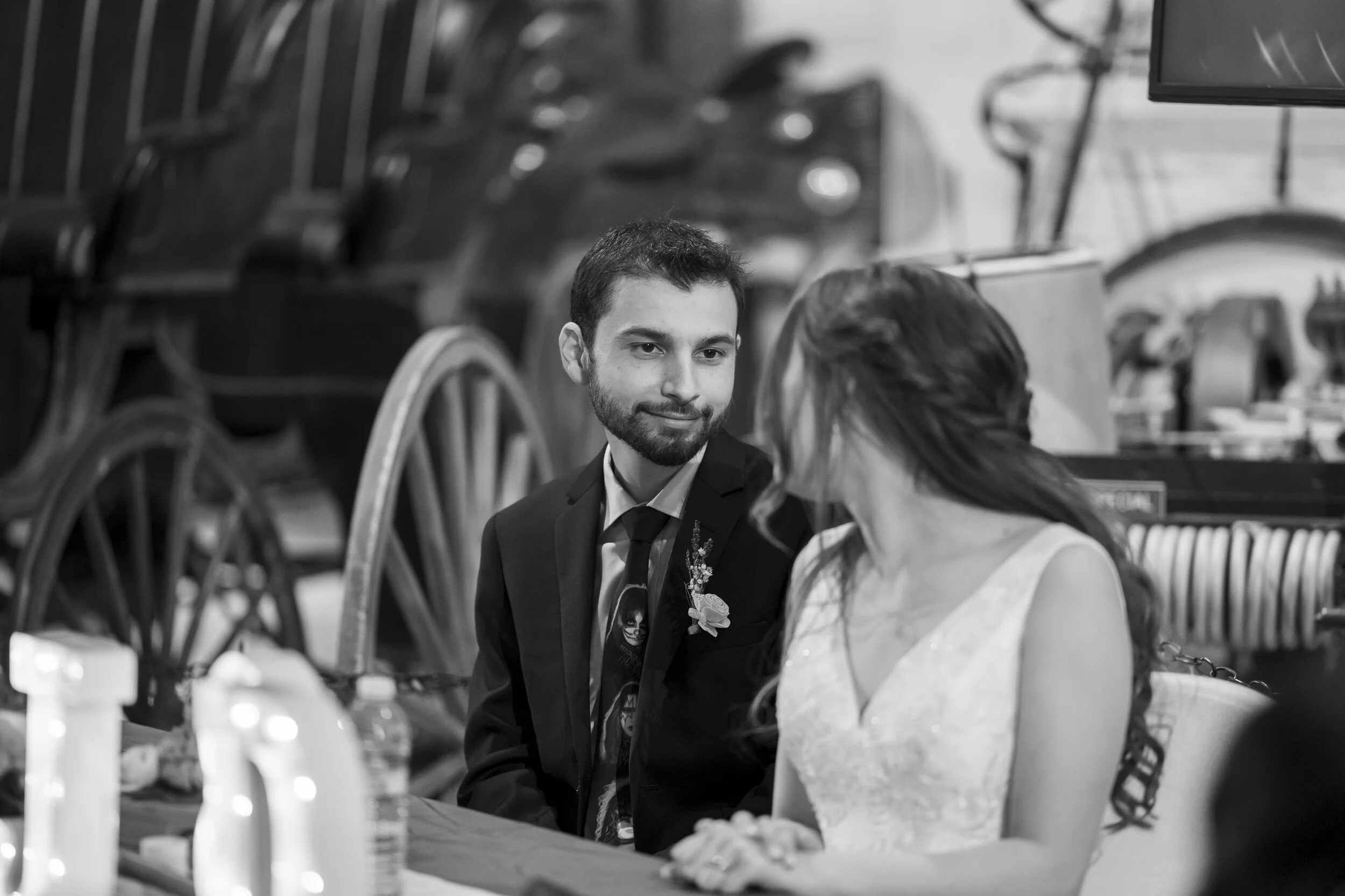 A man in a suit and a woman in a wedding dress sitting at a table in front of large train wheels, engaged in conversation. The setting appears to be indoors.