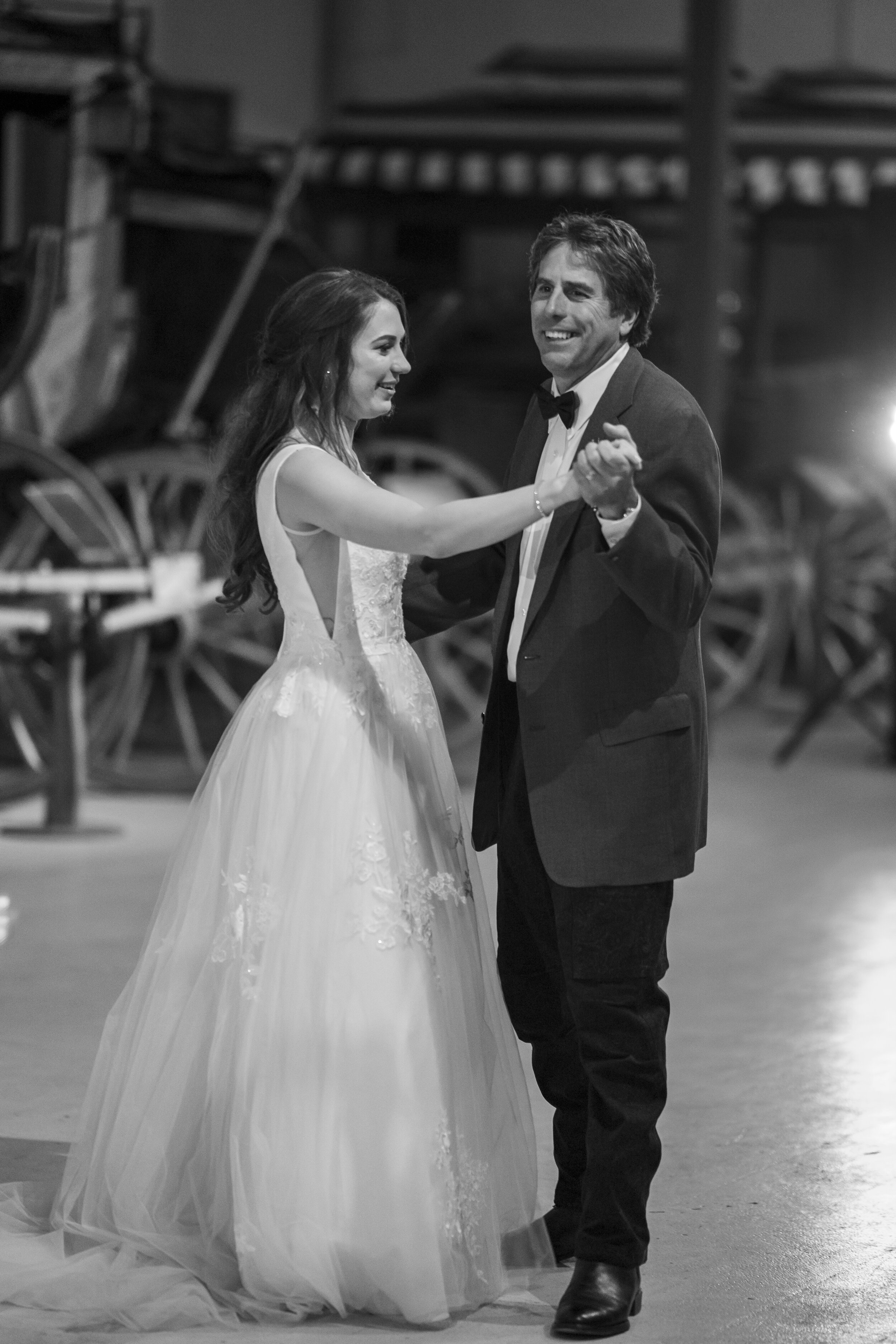 A black and white photo of a young woman in a wedding dress dancing with a man in a tuxedo, both smiling, in a rustic setting with wooden wagons in the background.