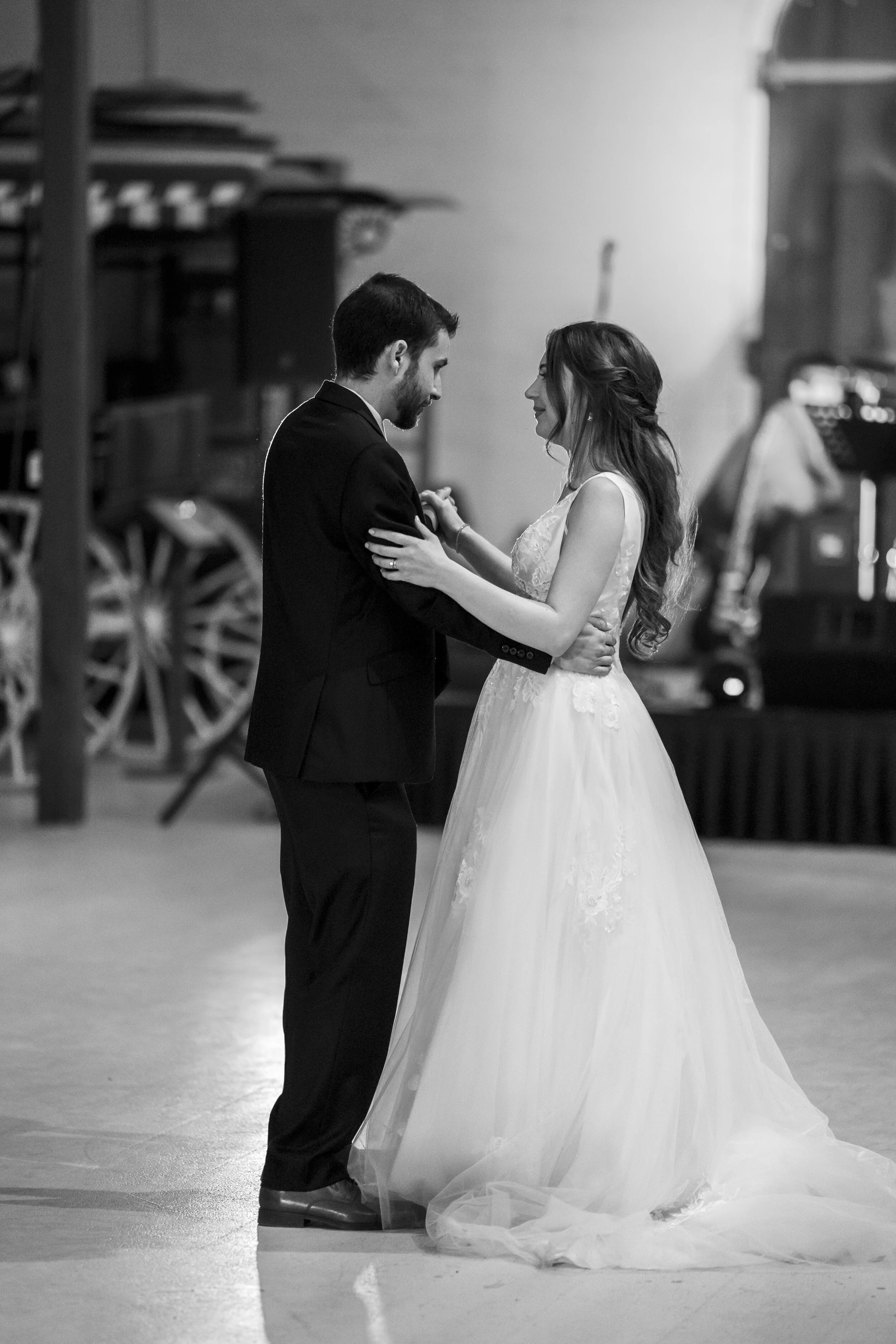 A black and white photo of a bride and groom dancing in a ballroom or event space. The bride is wearing a wedding dress with a long train, and the groom is in a suit. They are facing each other, holding hands, and smiling. In the background, there ar