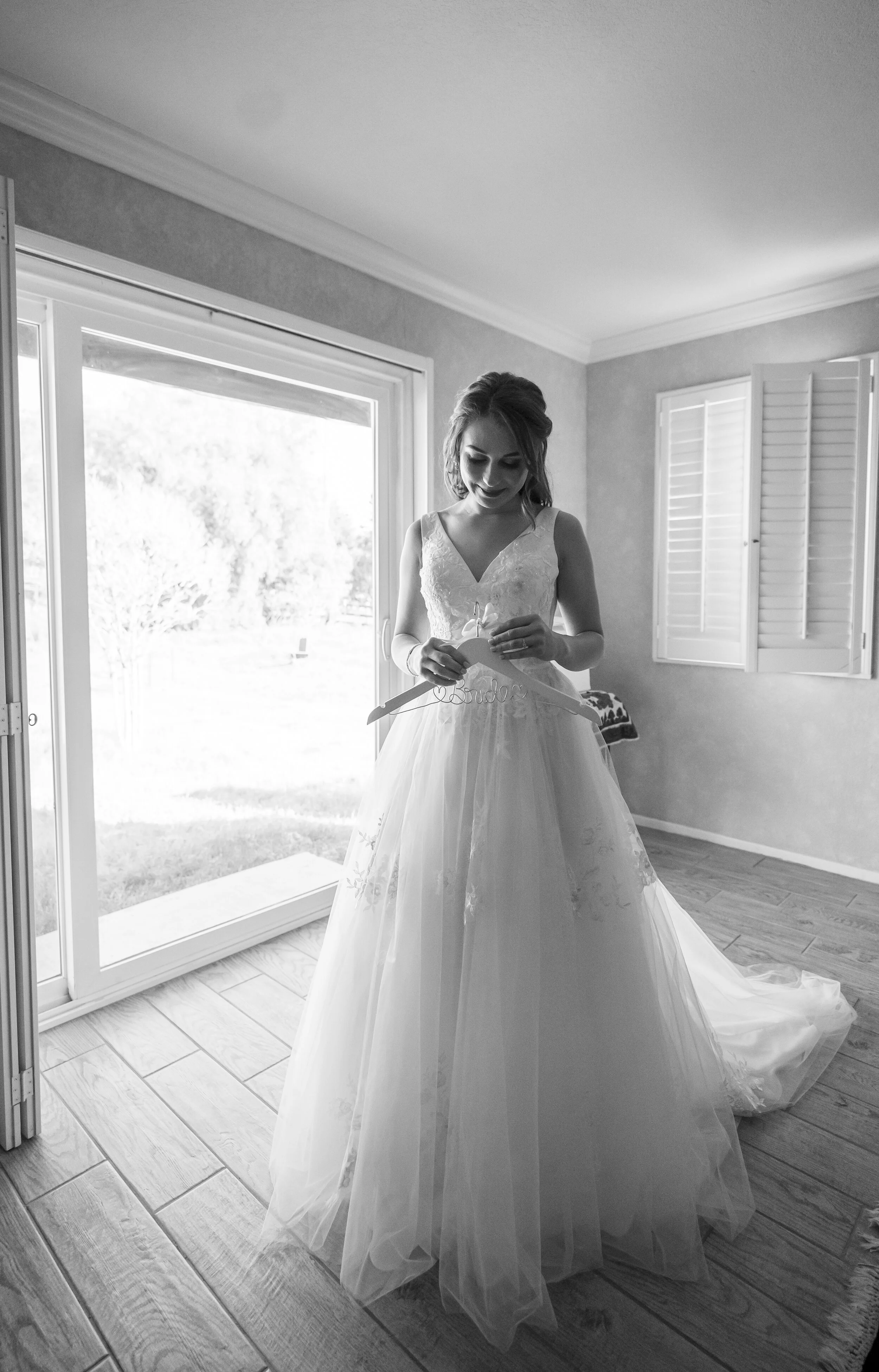 A woman in a wedding dress holding a wedding dress hanger labeled 'bride' standing indoors next to large sliding glass doors opening to a garden.