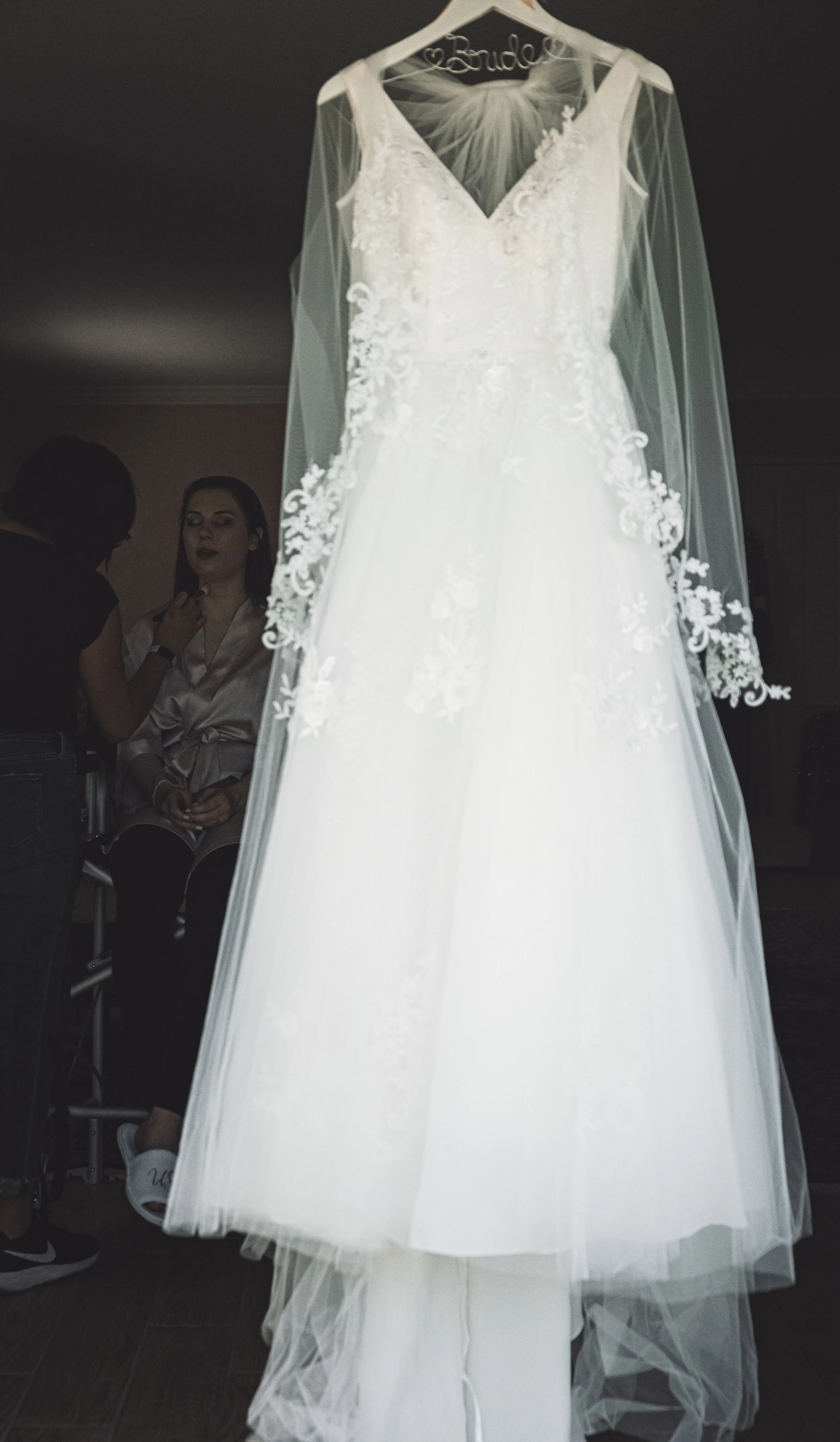 A white wedding dress with lace details hangs on a hanger in front of a mirror, with two women in the background preparing.