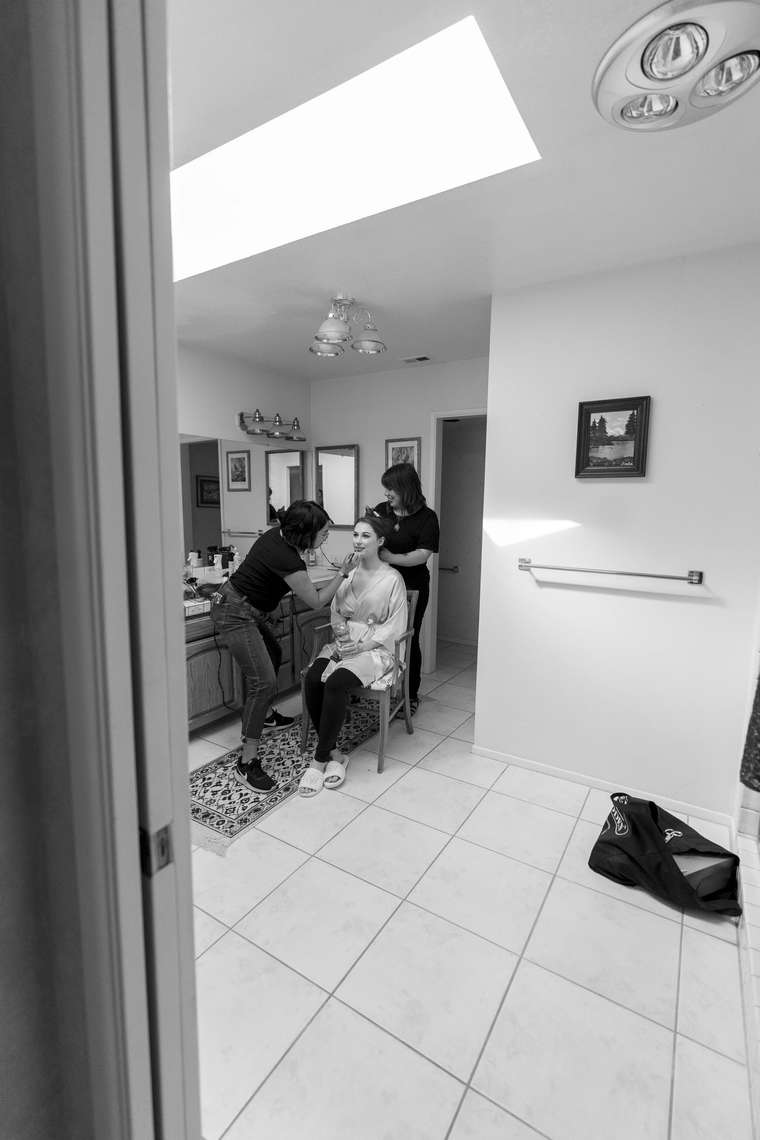 A woman sitting in a chair getting her makeup done by a makeup artist in a bathroom, while another woman looks on. The scene is viewed through a doorway, with framed pictures on the wall and a towel bar visible.