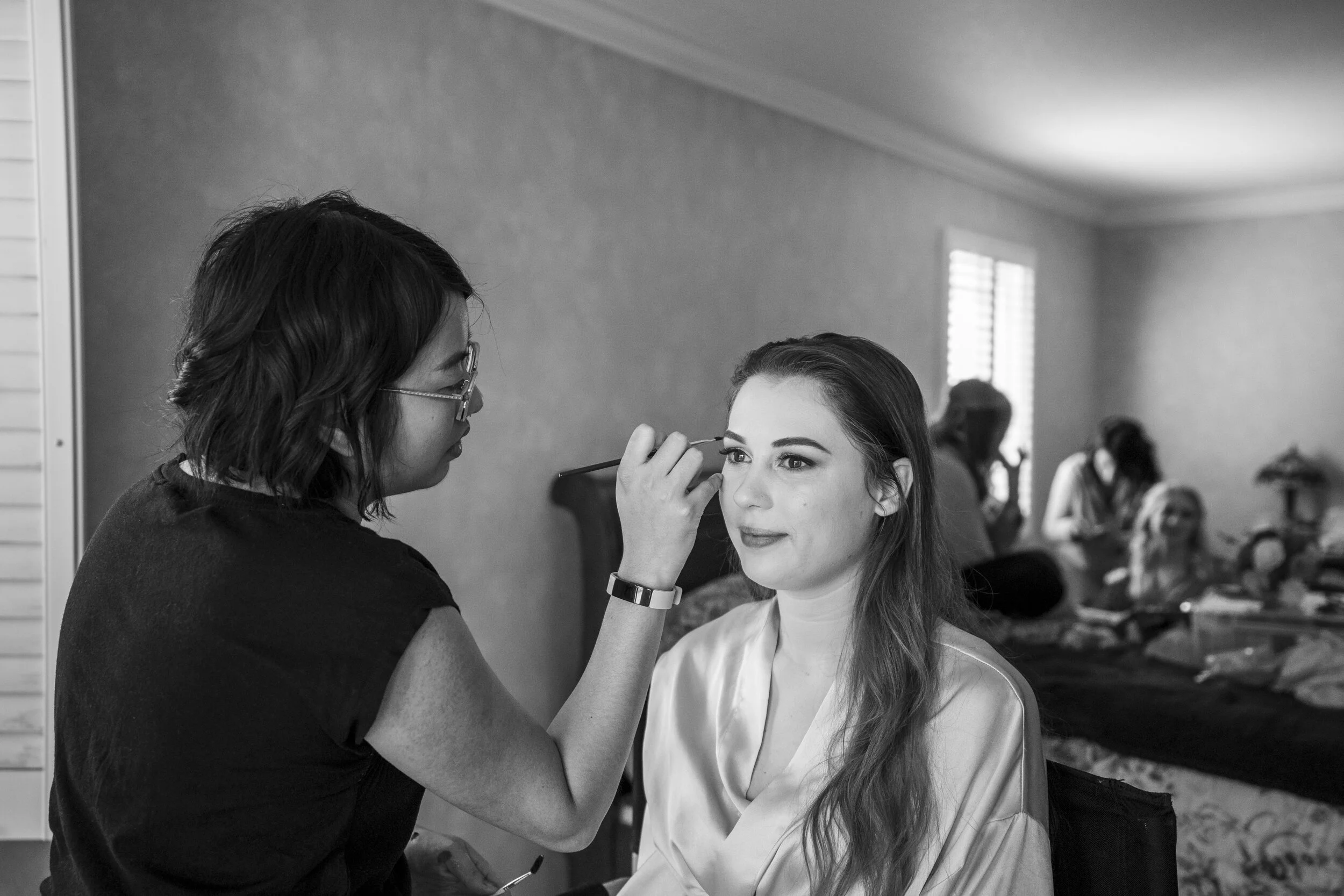 A woman is receiving makeup application from a makeup artist inside a well-lit room with multiple people seated in the background.
