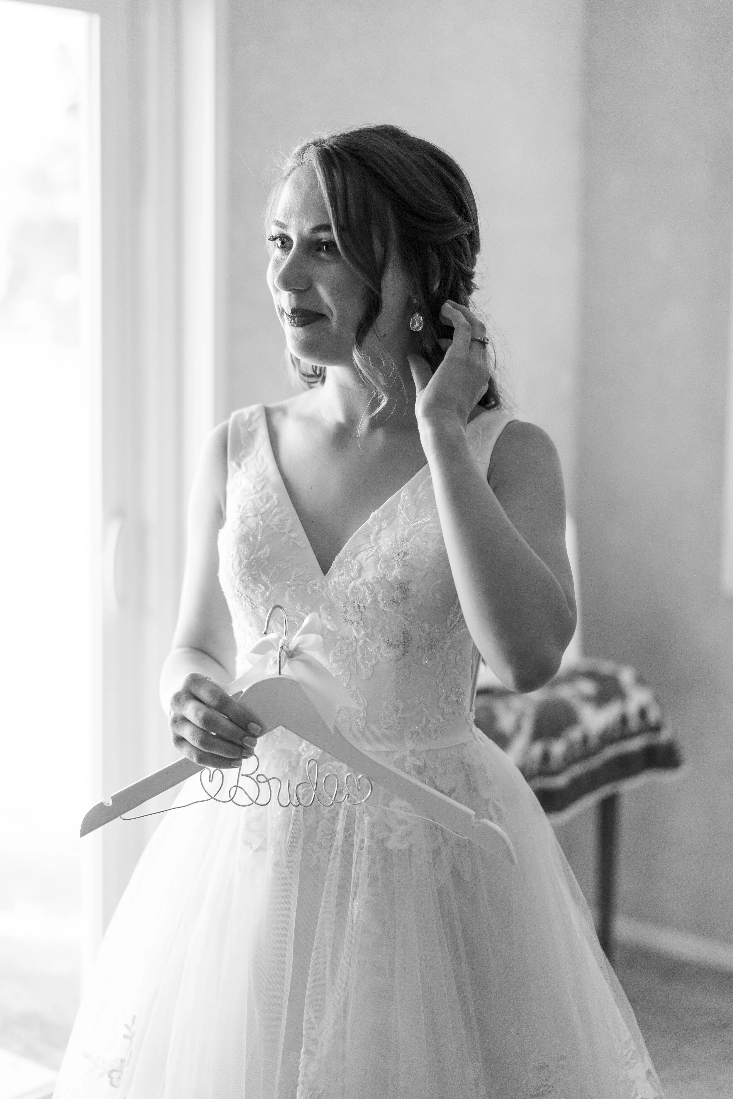 A bride dressed in a wedding gown, holding a hanger with the word 'bride' on it, standing near a window with soft natural light.