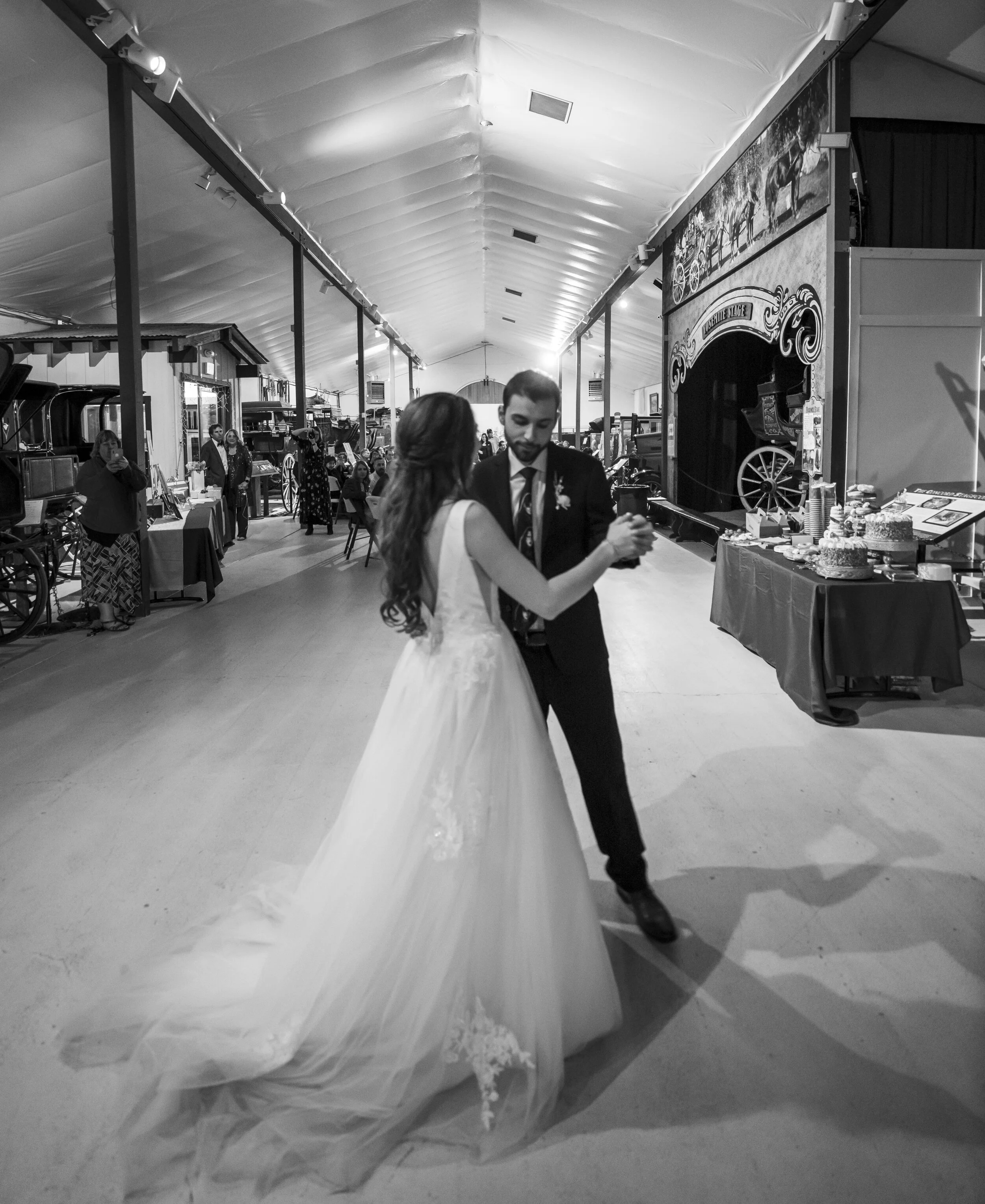 A wedding reception with a bride and groom dancing in a large tent. The tent has a high, padded ceiling, and there are vintage carriages and various display booths along the sides. A table with cake and refreshments is visible on the right. The scene