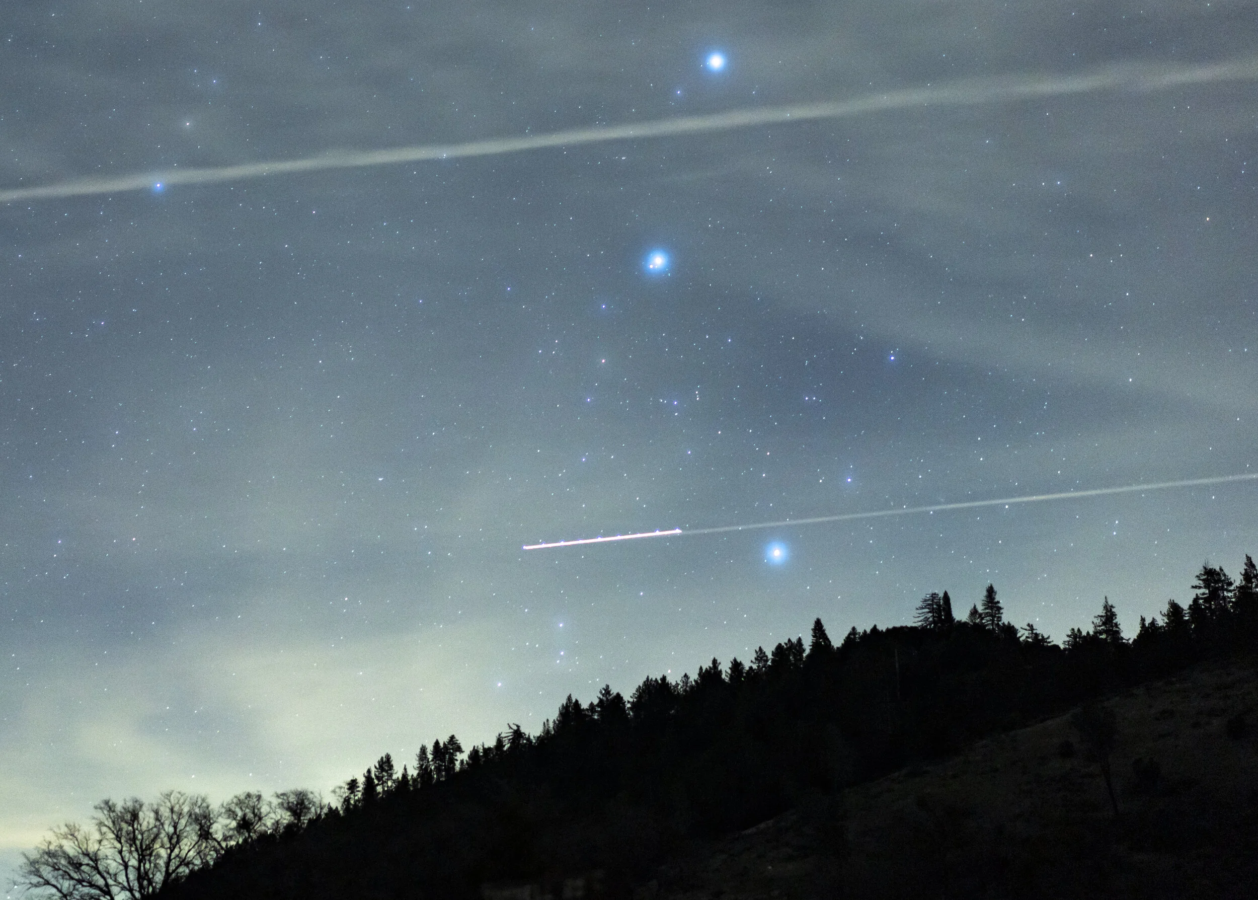 Night sky filled with numerous stars, with a few bright objects and thin, wispy clouds. Silhouetted trees and hills are visible at the bottom of the image.