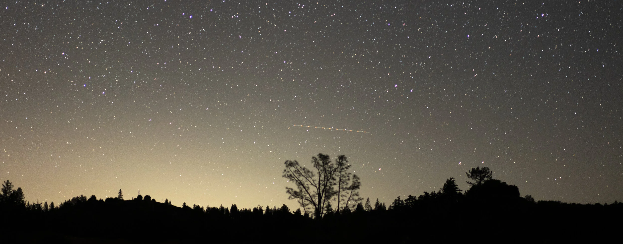Night sky filled with stars over a silhouette of trees and hills, with a faint line of clouds and a meteor streaking across.