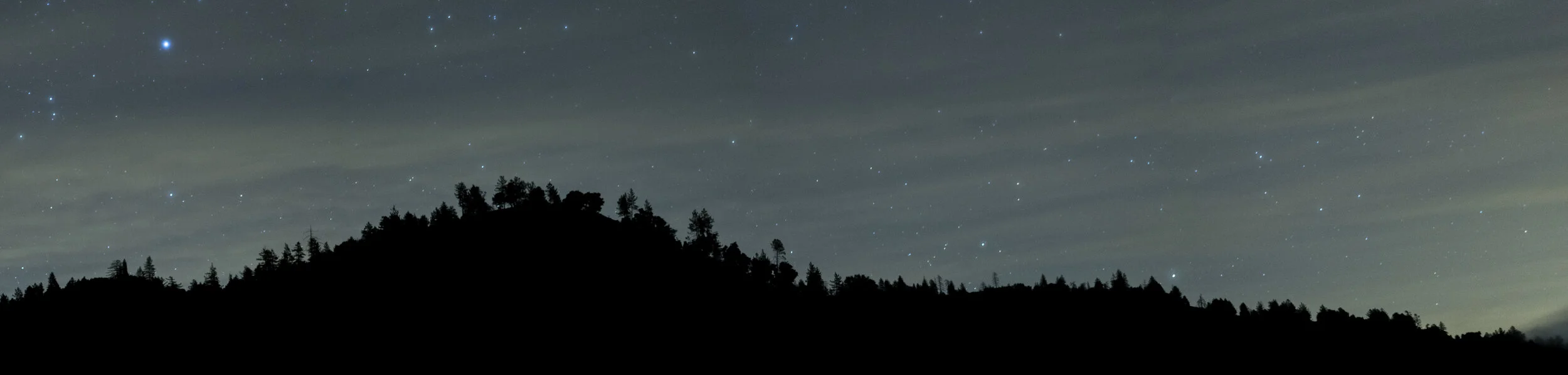 Night sky with stars above a silhouette of a forested hill or mountain.