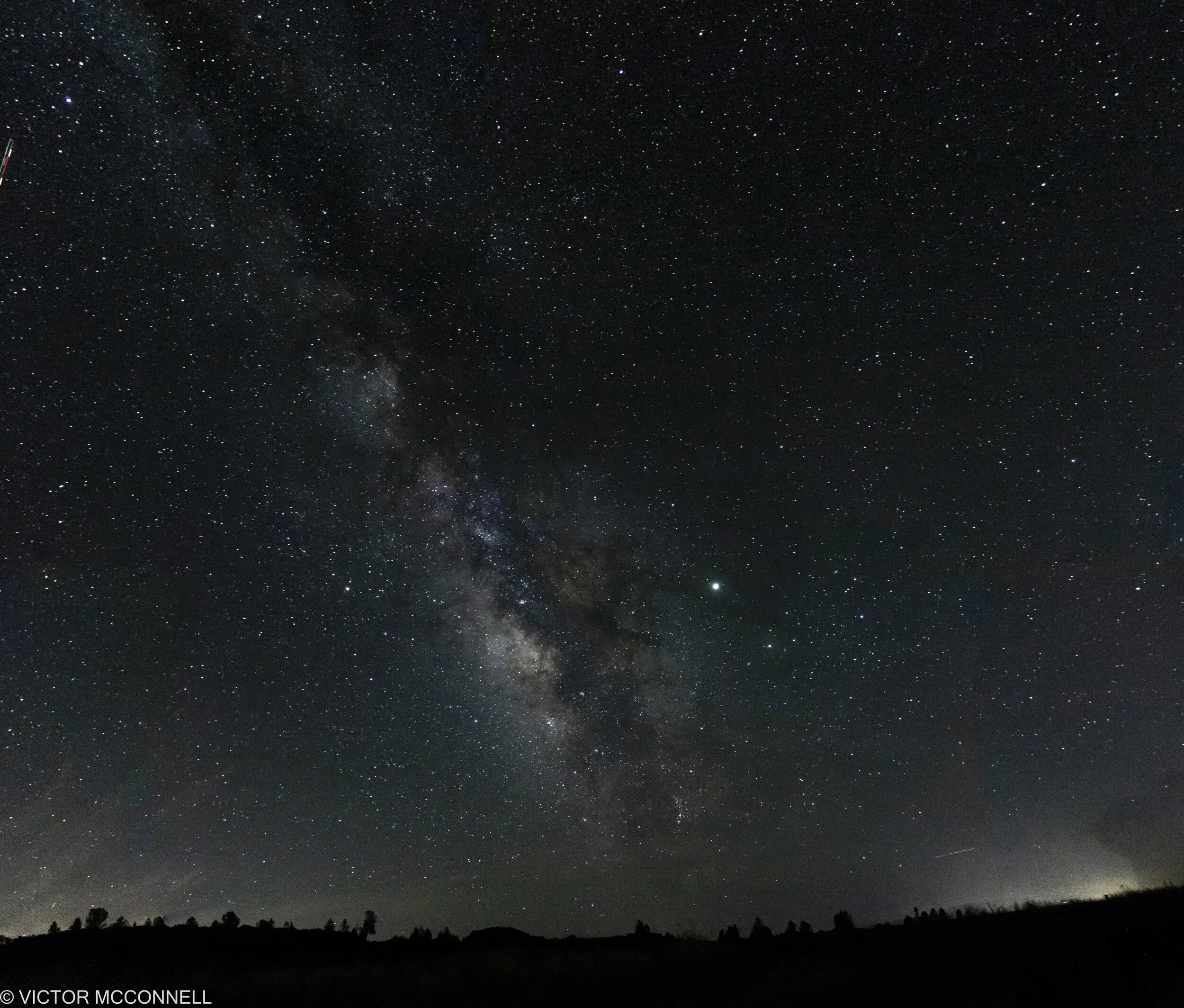 A wide view of a starry night sky with the Milky Way galaxy clearly visible, over a silhouette of a treeline.