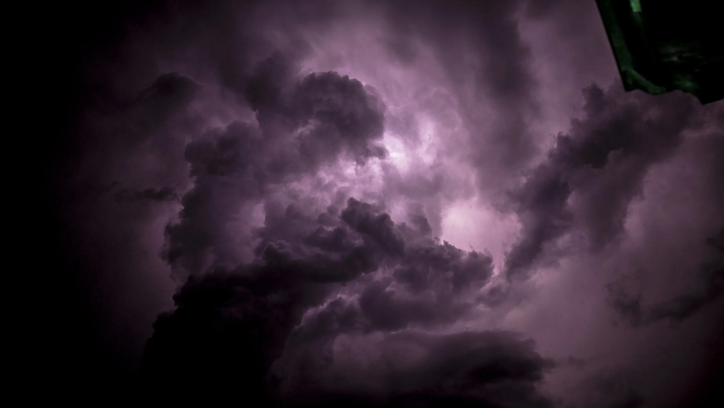 Dark storm clouds illuminated by lightning with purple hues, viewed from below.