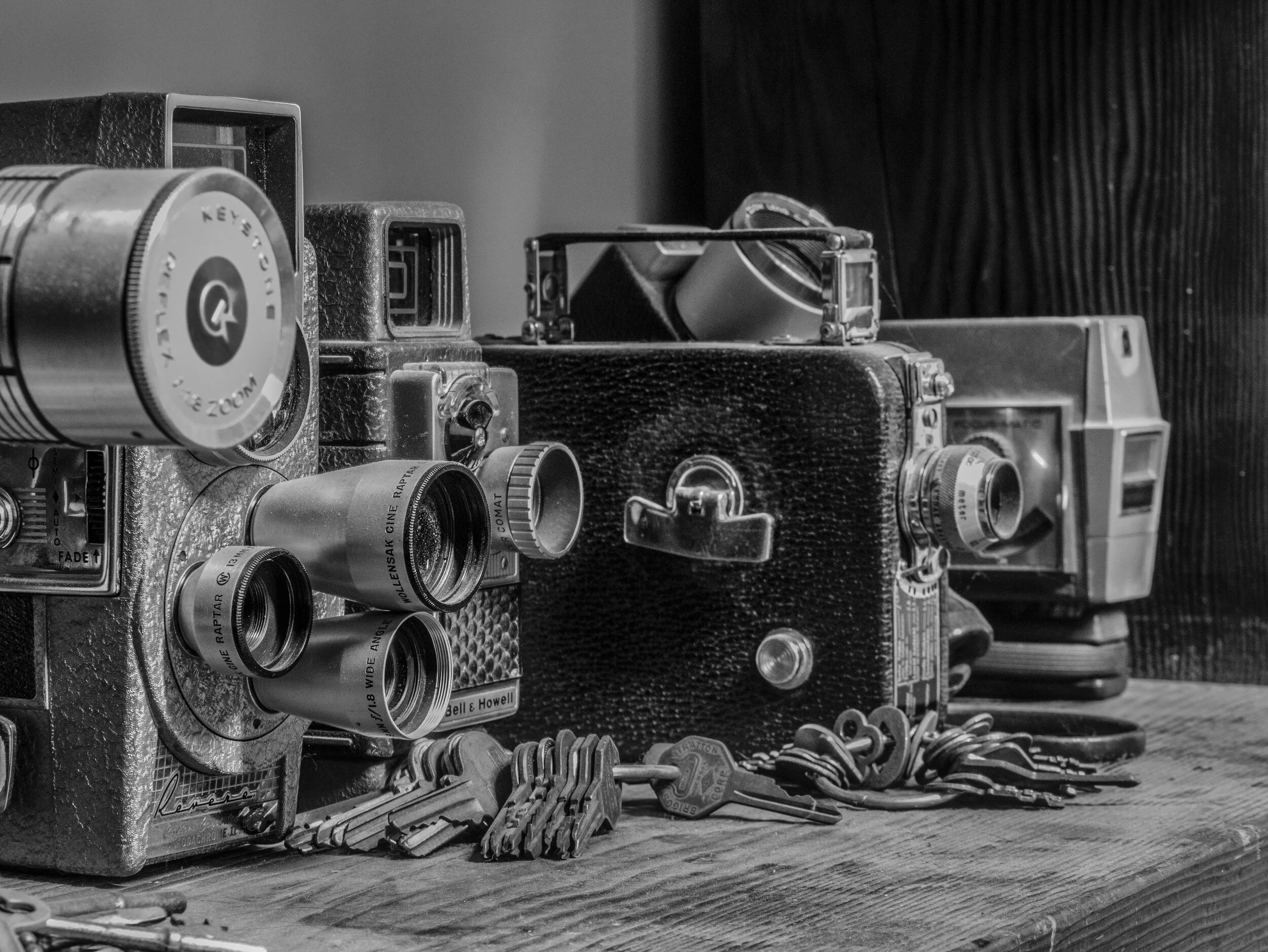 Black and white photo of vintage cameras, lenses, and camera accessories arranged on a wooden surface.