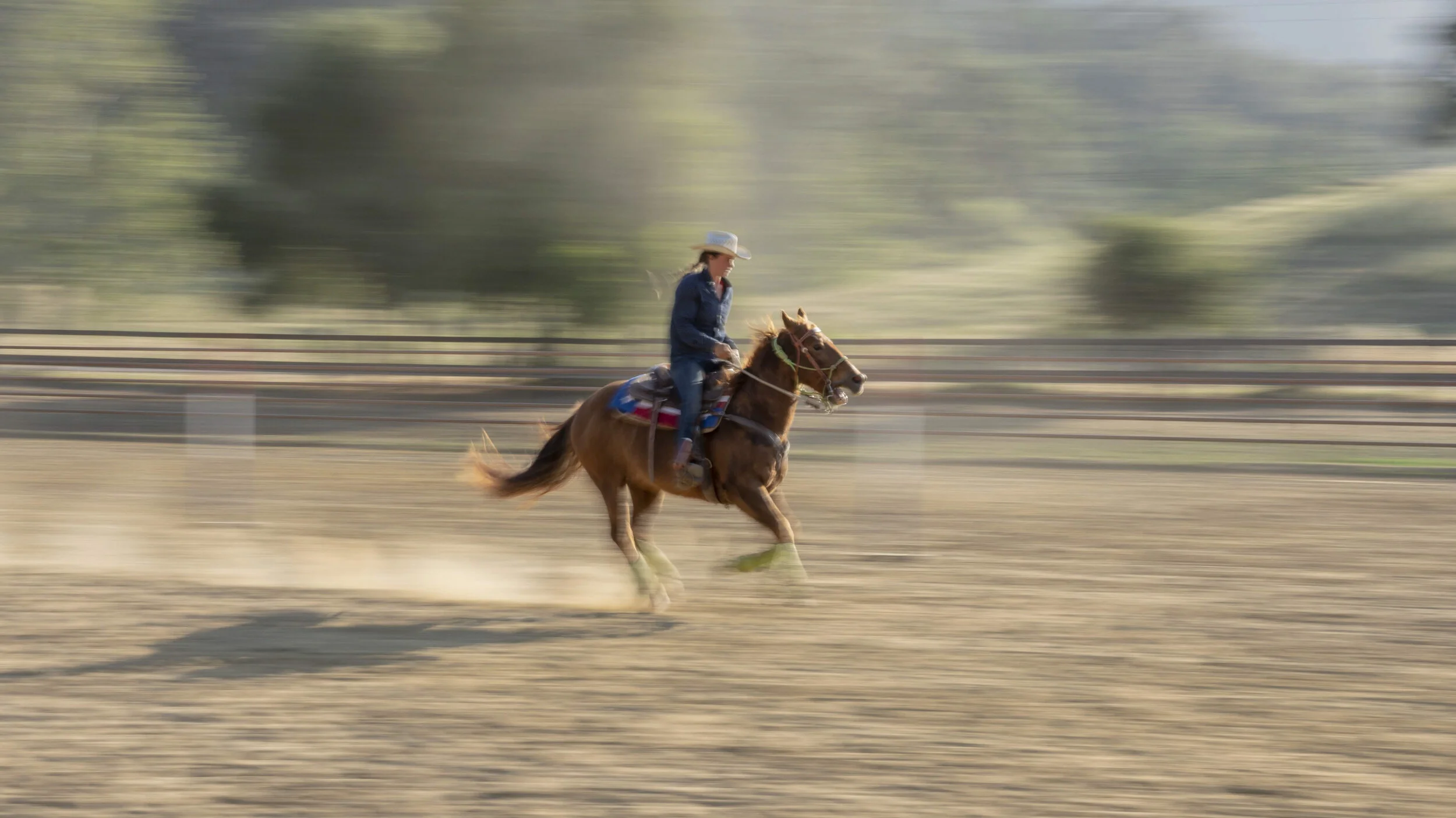 A woman riding a galloping horse on a dirt track with a blurred background, wearing a cowboy hat and denim jacket.