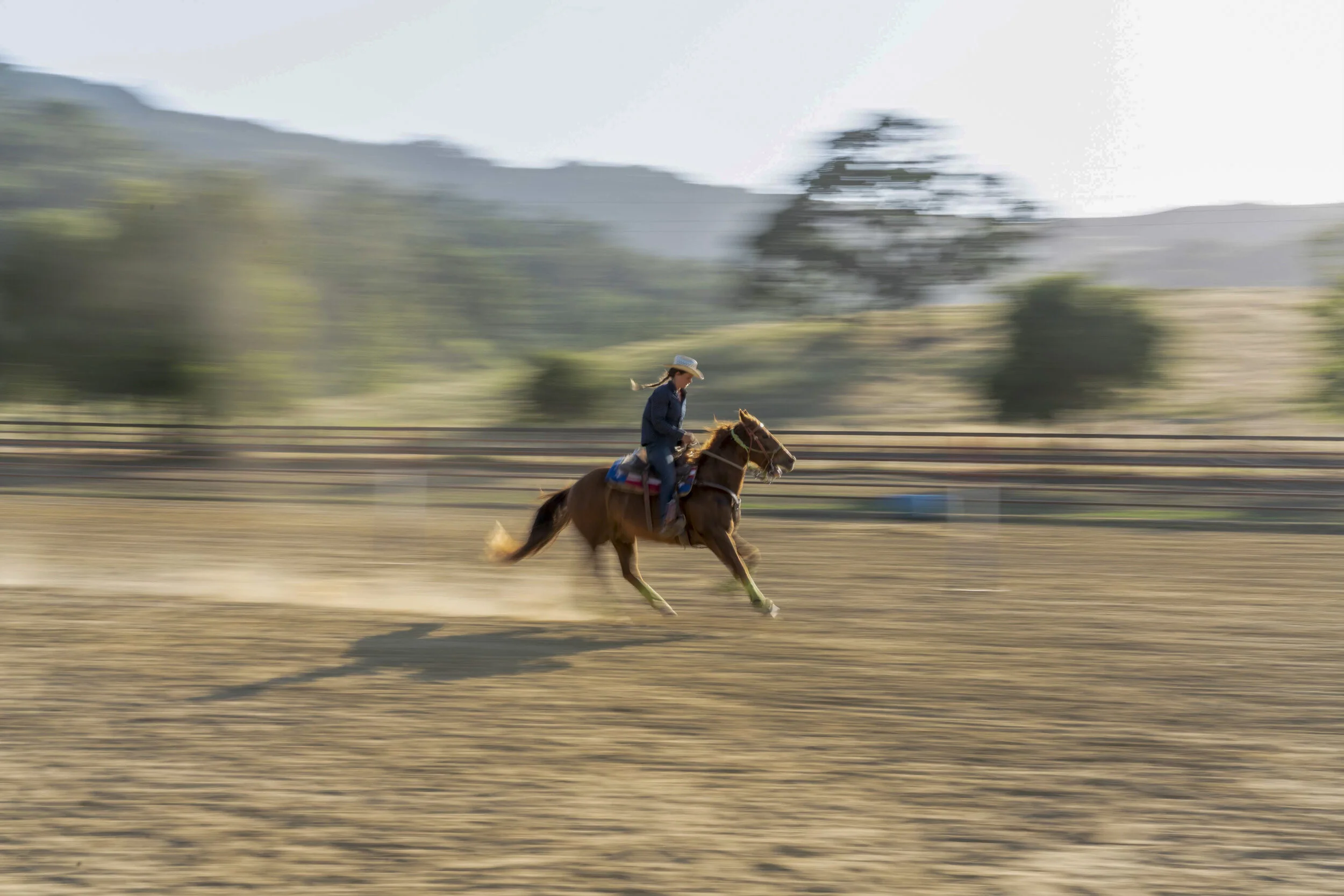 A woman wearing a cowboy hat riding a galloping brown horse on an open field with trees and hills in the background.