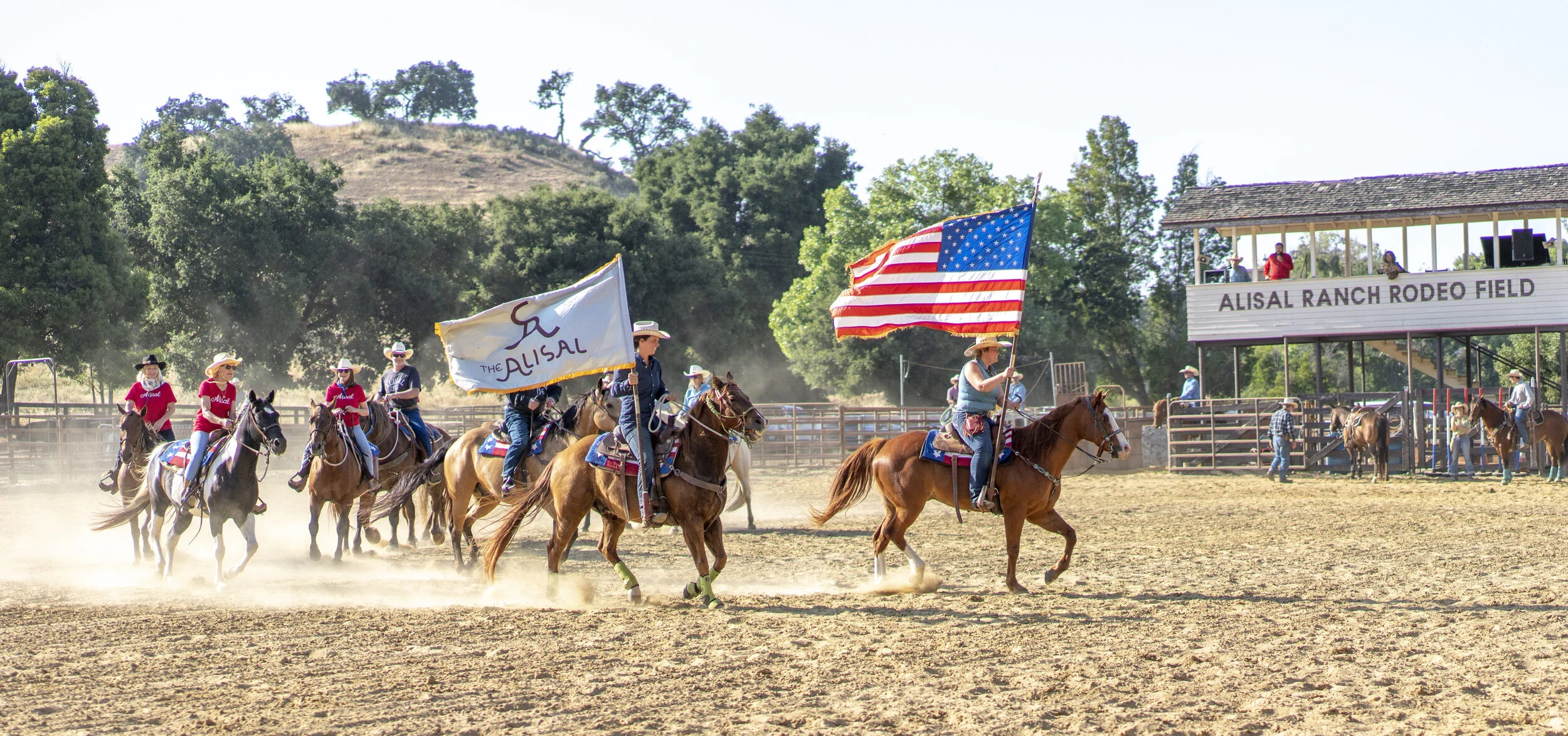 People riding horses in a rodeo arena holding American flags, with a building labeled "Alisal Ranch Rodeo Field" in the background.
